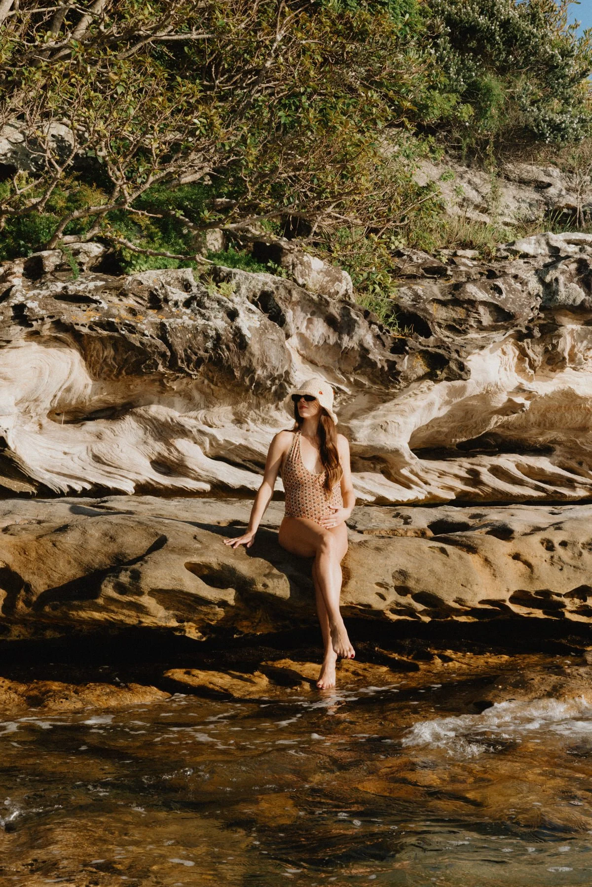 A woman in a patterned swimsuit and hat sits on a large rock by a rocky shoreline with green bushes and trees in the background.