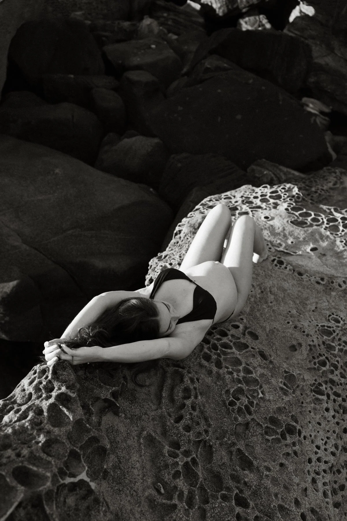 A woman lying on a rocky beach with her arms behind her head, wearing a black swimsuit and a lace dress. The scene is in black and white.