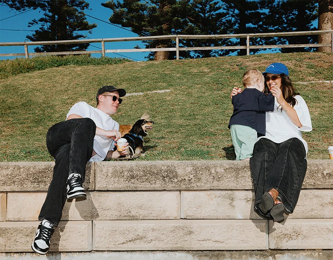 A young man sitting on a stone ledge with a dog, holding a drink, and a woman with a kid, smiling and interacting, outdoors on a grassy hill with trees and a fence in the background.