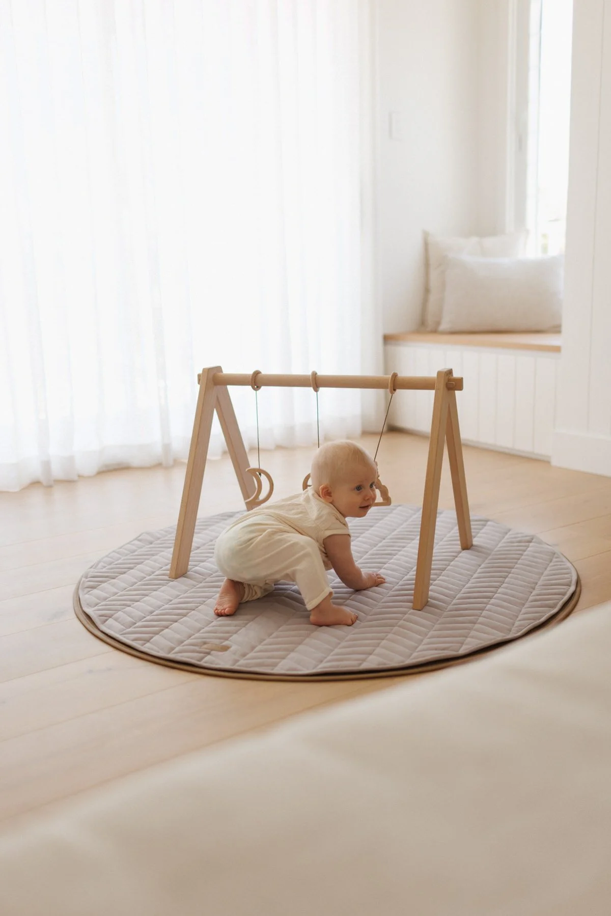 A baby crawling on a padded circular rug on a wooden floor, beneath a wooden baby gym with rings. The room has white curtains, a window seat with a cushion, and soft natural lighting.