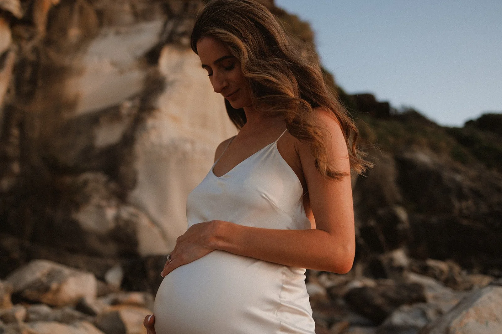 Pregnant woman wearing white silk dress against a rocky backdrop