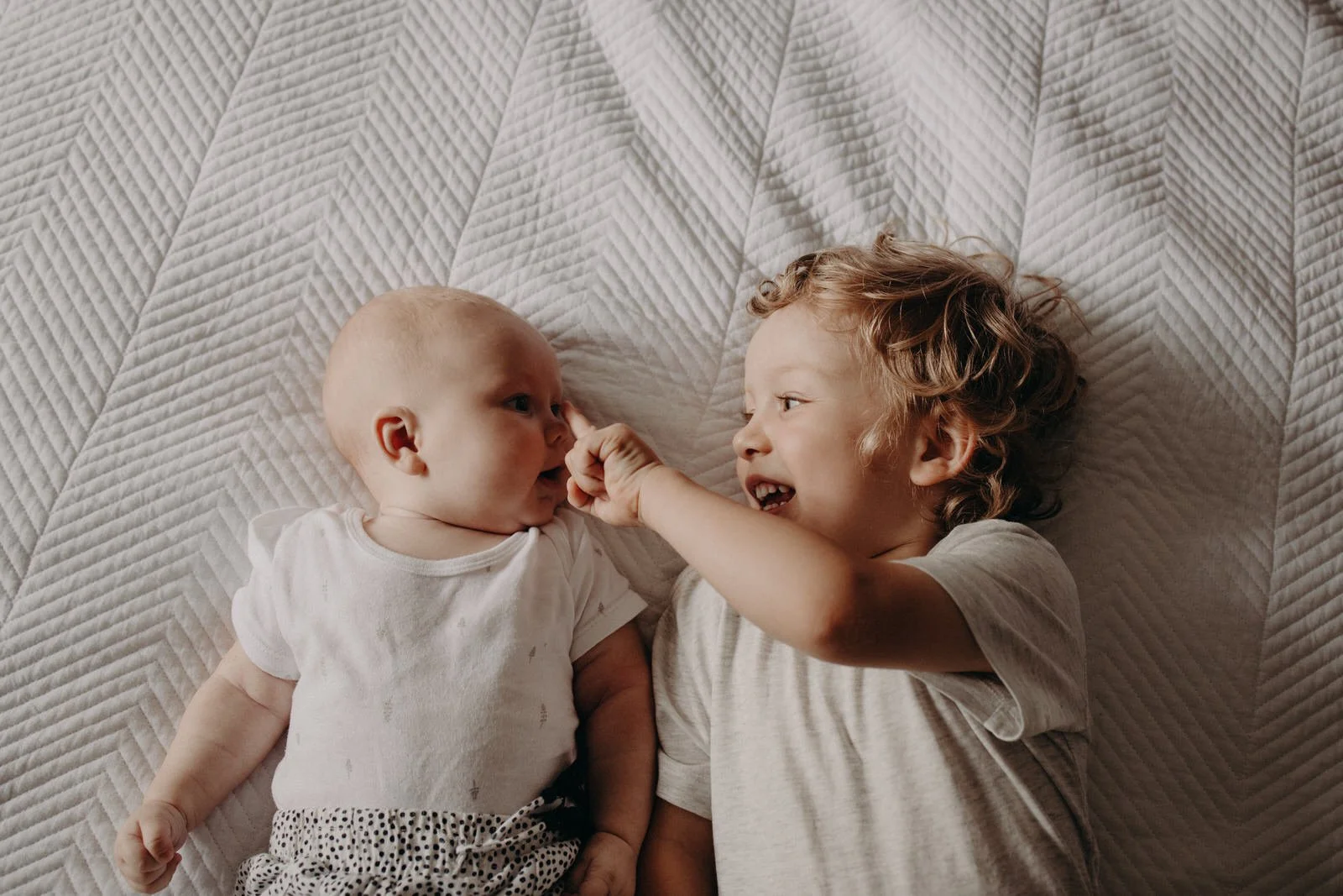 A photo of a toddler brother with his baby sister on a bed