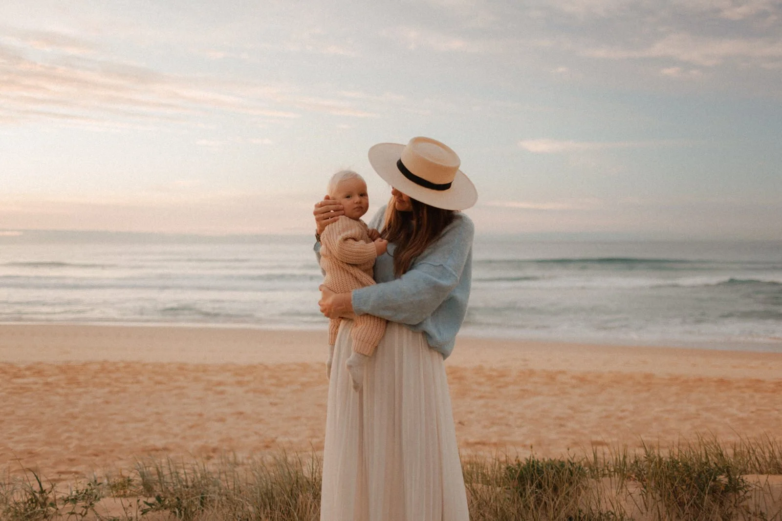 A woman in a hat holds a child on a sandy beach, with the ocean and a pastel sky in the background.