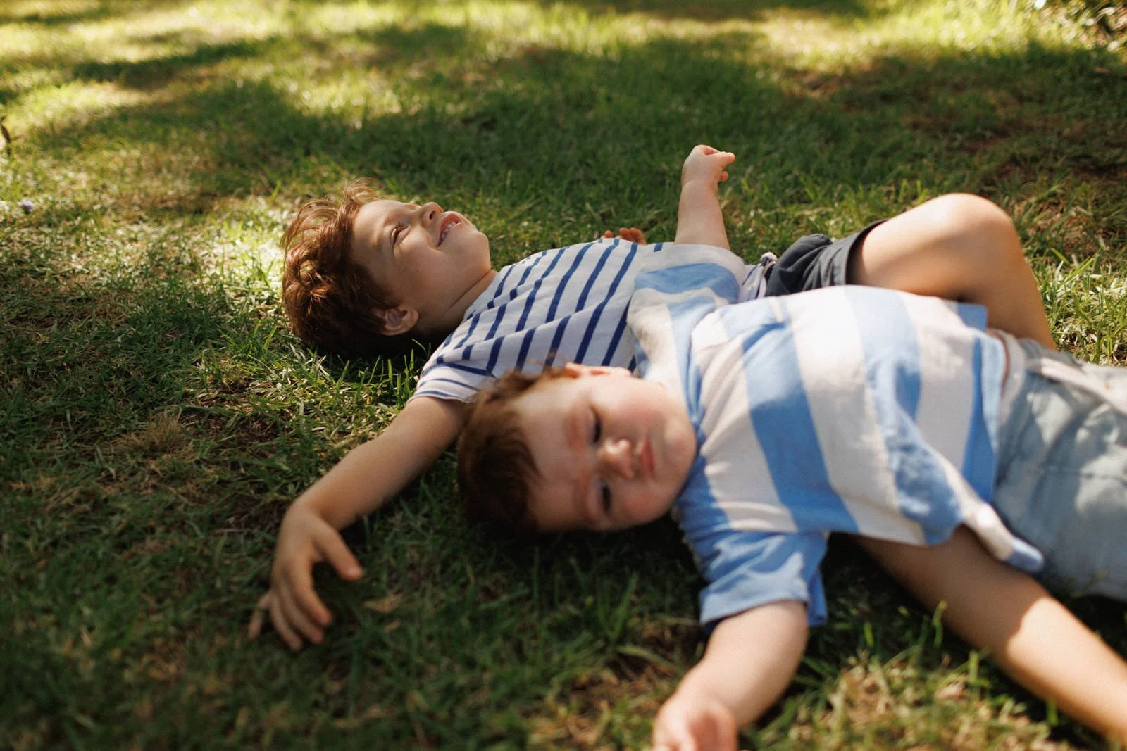 Two young boys in striped shirts lie on the grass, relaxing in the dappled sunlight of a garden.