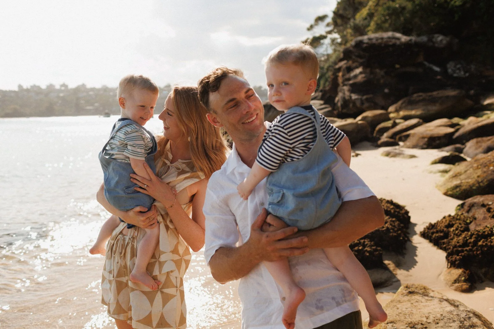 A smiling couple holds their twin toddlers on a sunny beach, with water sparkling in the background.