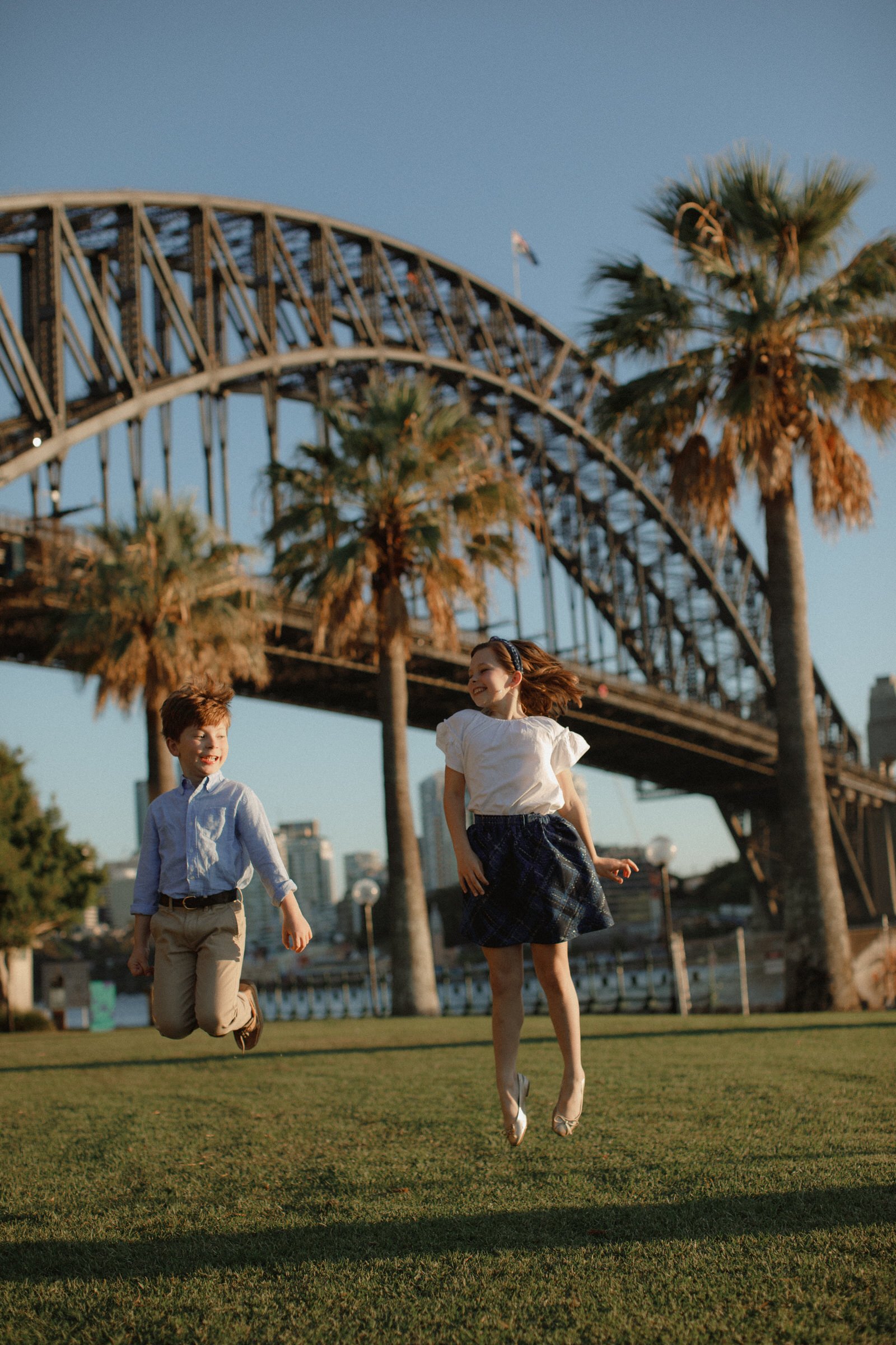 Two children jump playfully on a grassy area, with palm trees and the Sydney Harbour Bridge in the background.
