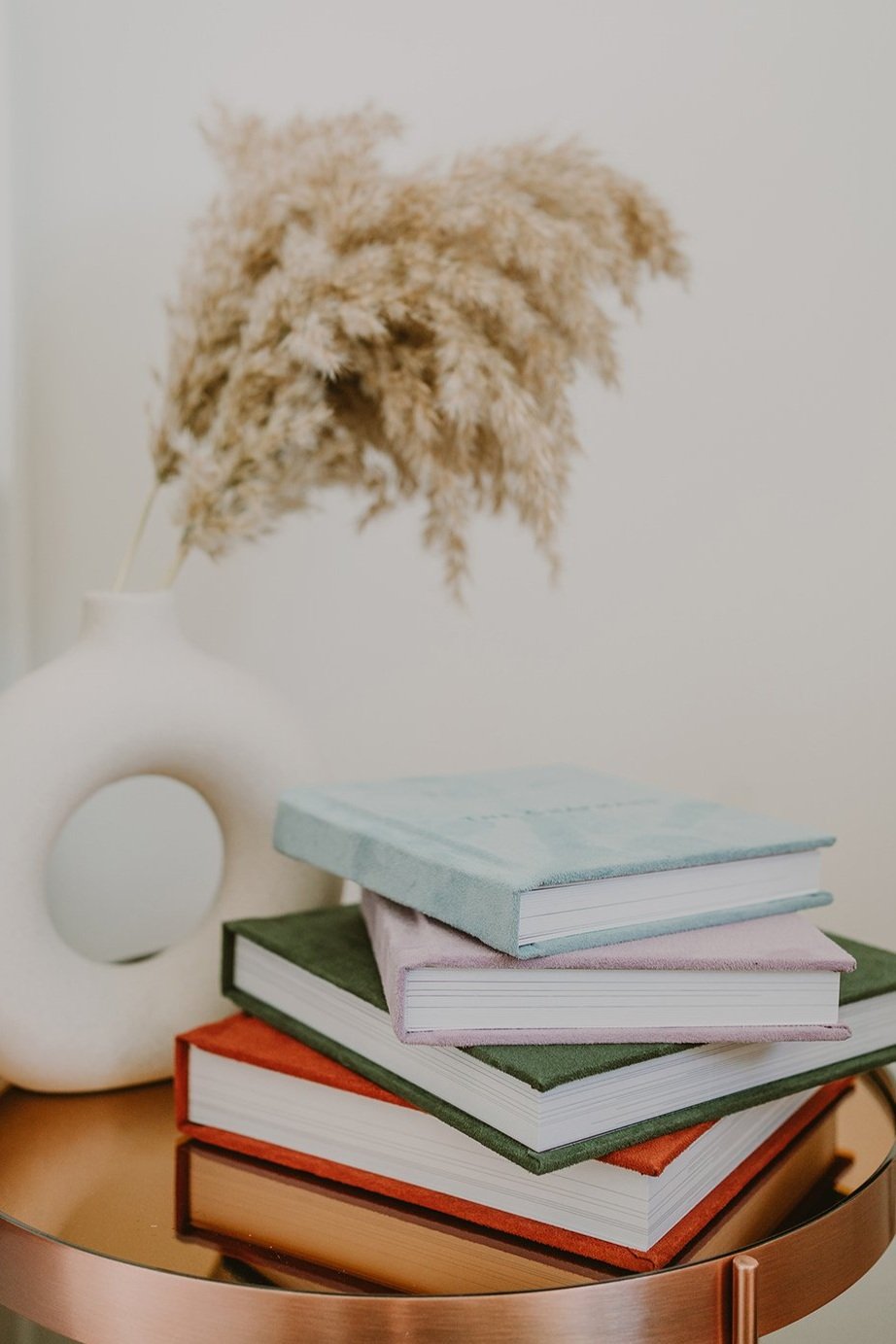 Stack of four hardcover books with pastel-colored covers, placed on a round side table. Behind the books is a decorative white ceramic vase with a circular cutout, holding beige dried pampas grass.