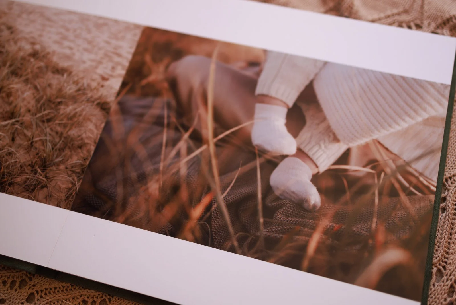 Photograph of a person's legs wearing white knit socks and beige pants, resting on a brown textured blanket with dried grass or straw scattered around.