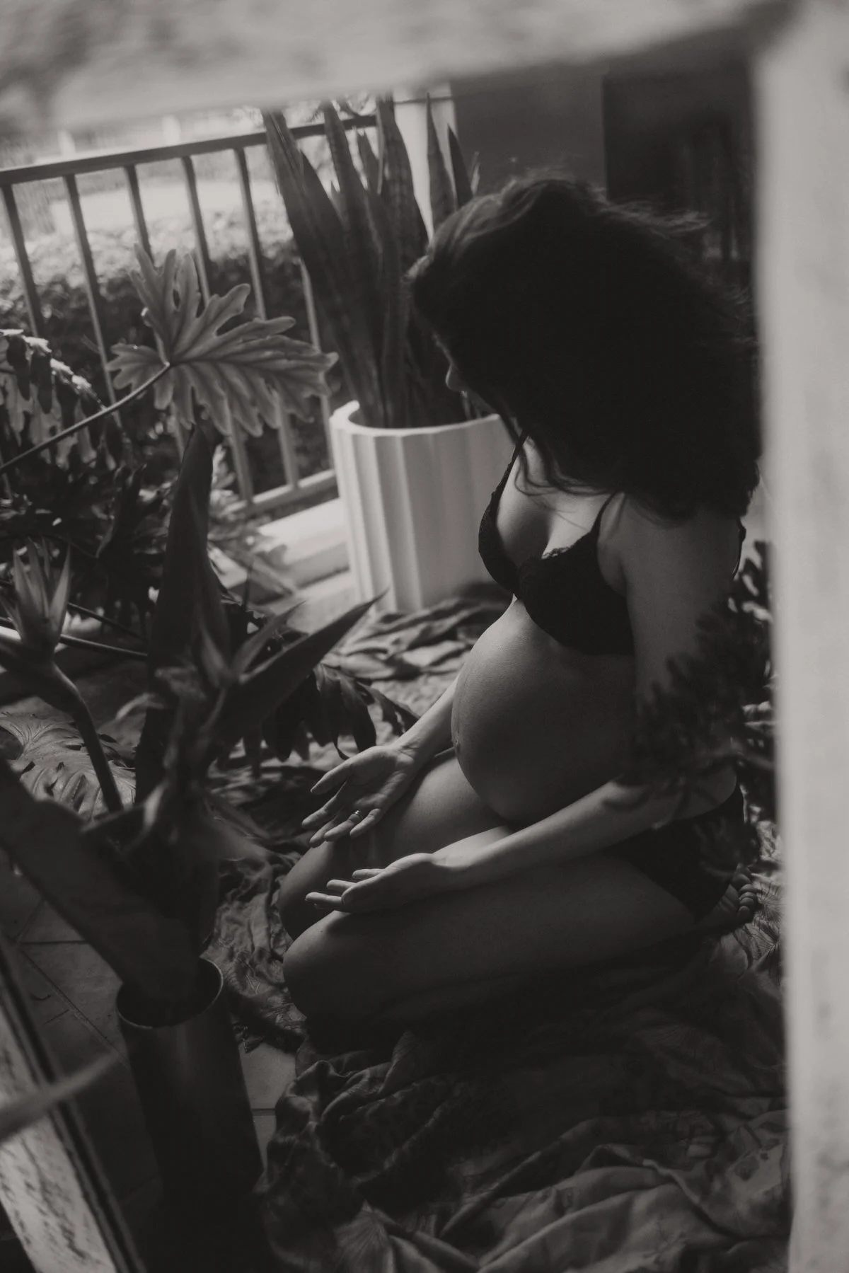 A woman sitting on the floor near a balcony or window with large plants nearby. She is wearing black lingerie and has long dark hair. The photo is black and white.