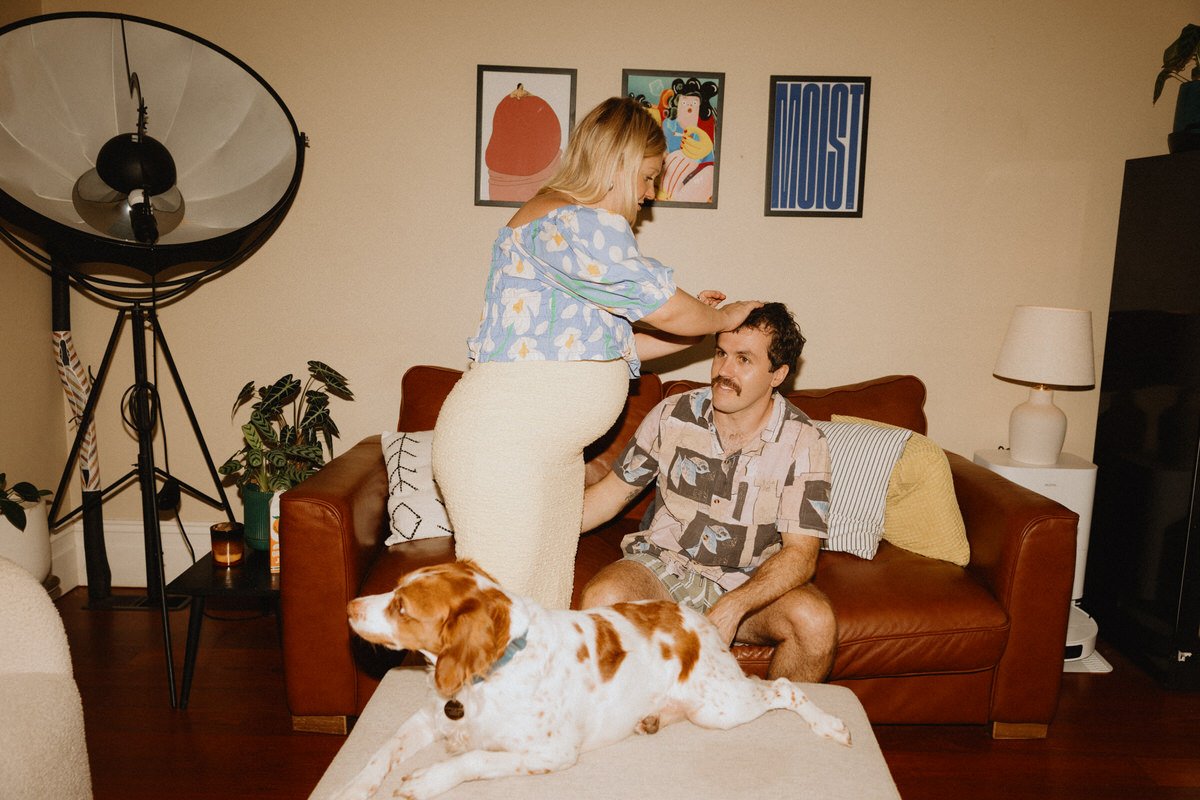 A woman standing close to a man sitting on a brown leather sofa, with her hand on his head, in a cozy living room. A dog lies on a white ottoman in front of the sofa. The background features wall art, a lamp, and potted plants.
