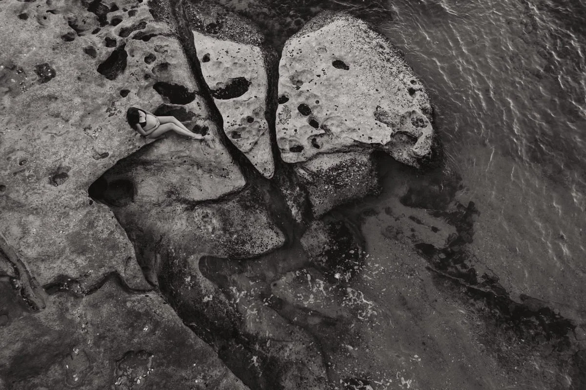A woman sitting on a large rock formation near the water's edge at the beach, taken from an aerial perspective in black and white.