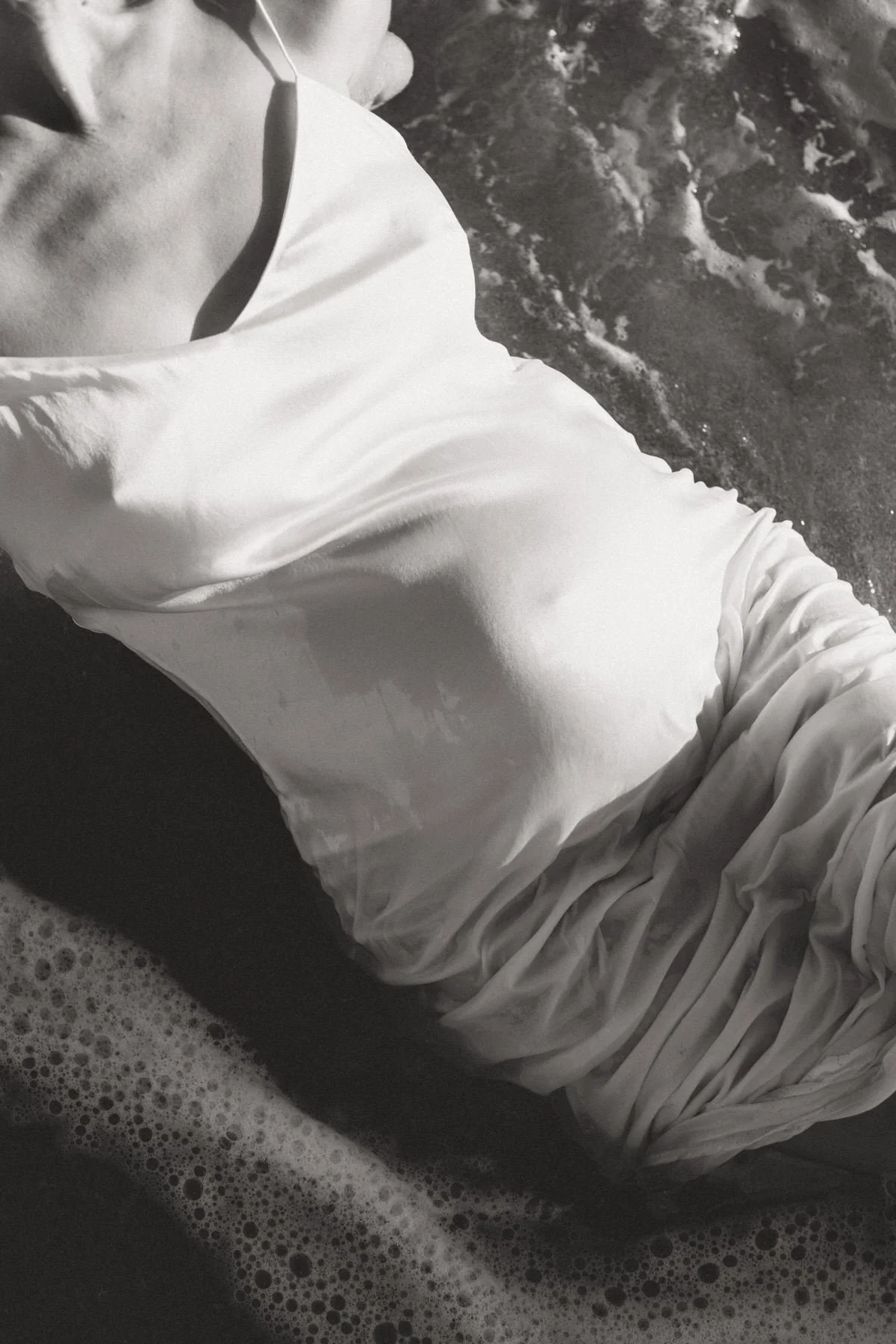 A person wearing a wet, white, long-sleeved shirt at the edge of the ocean, with waves washing over their arm and shoulder.