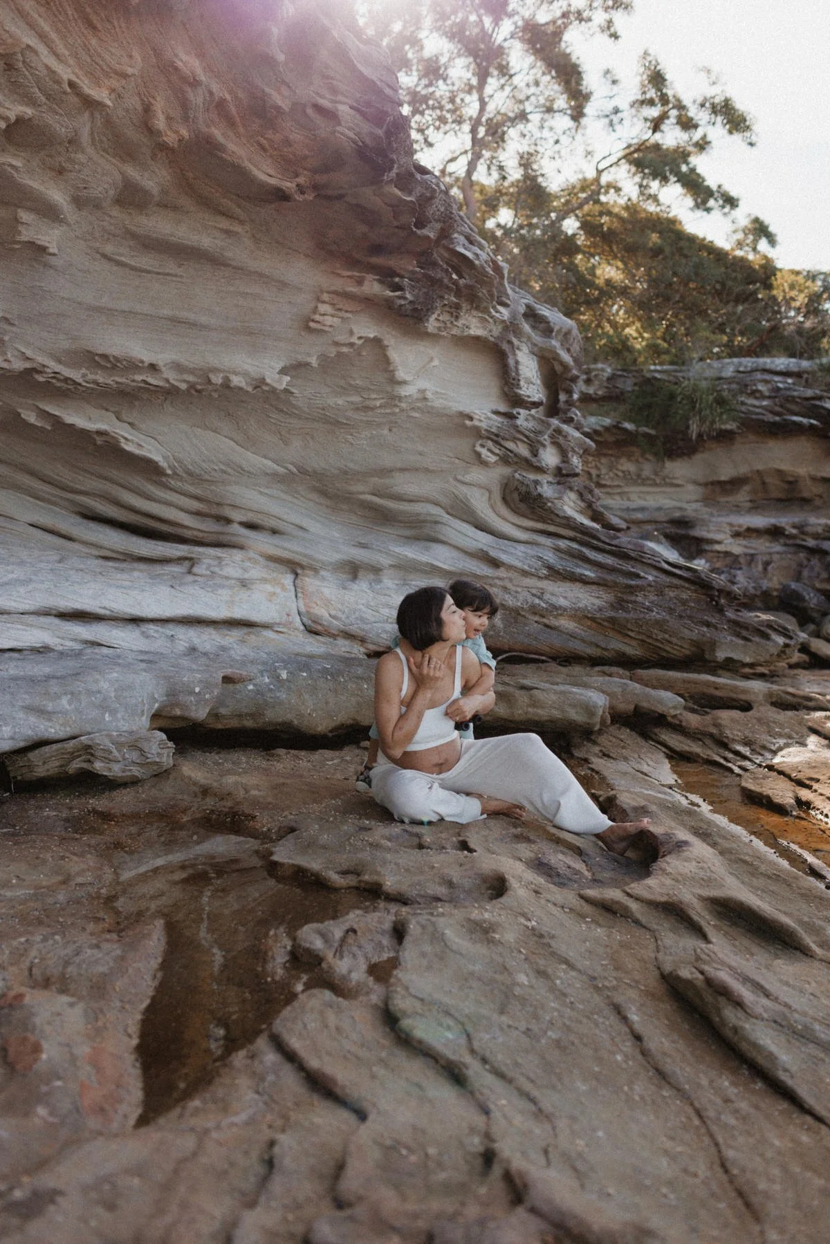 A woman and a young girl sitting on the rocks by a large sandstone cliff wall, with trees in the background during daytime.