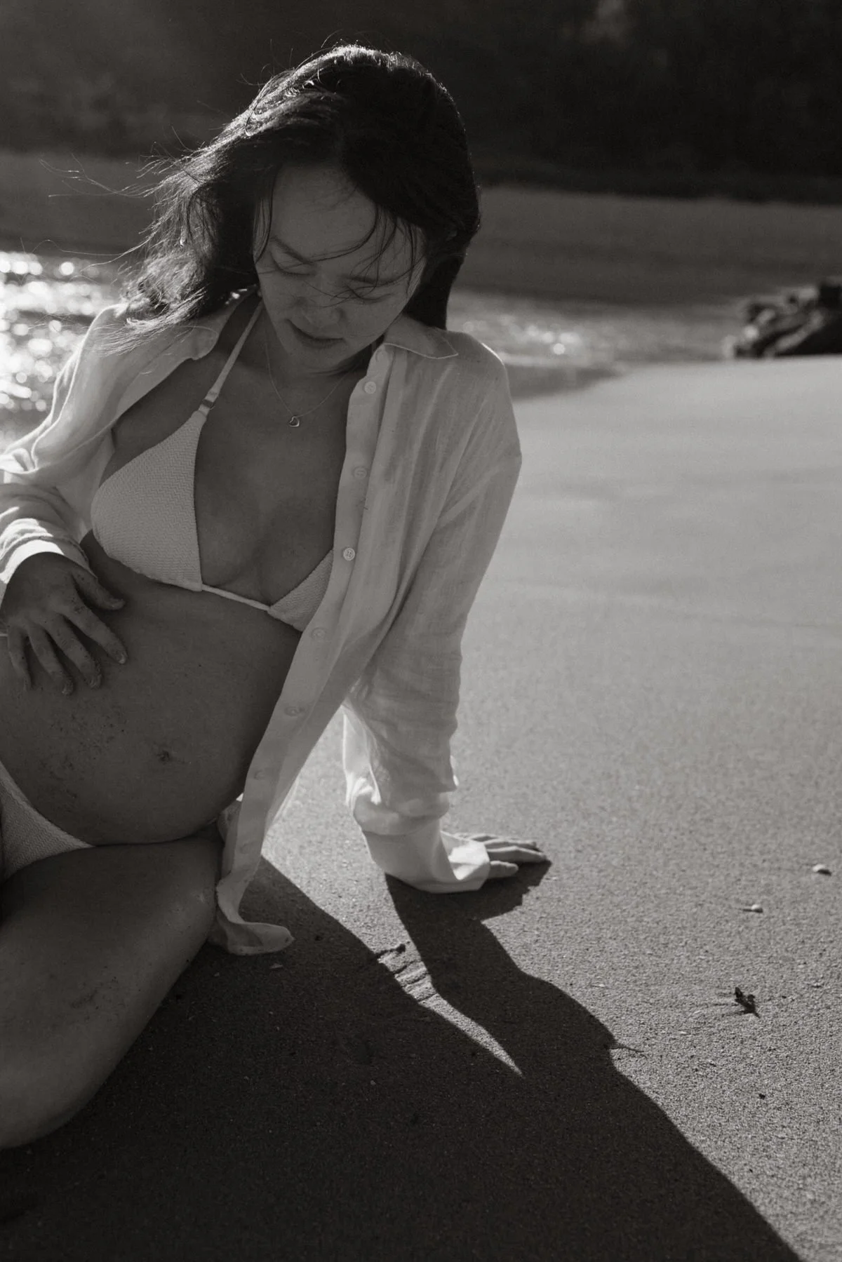 A woman on the beach with wind-blown hair, wearing an open shirt and a bikini, leaning on the sand with one hand, casting a shadow on the sand.