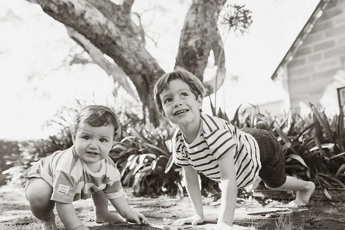 Two young children playing outdoors on the ground, crawling with trees and a house in the background, black and white photo