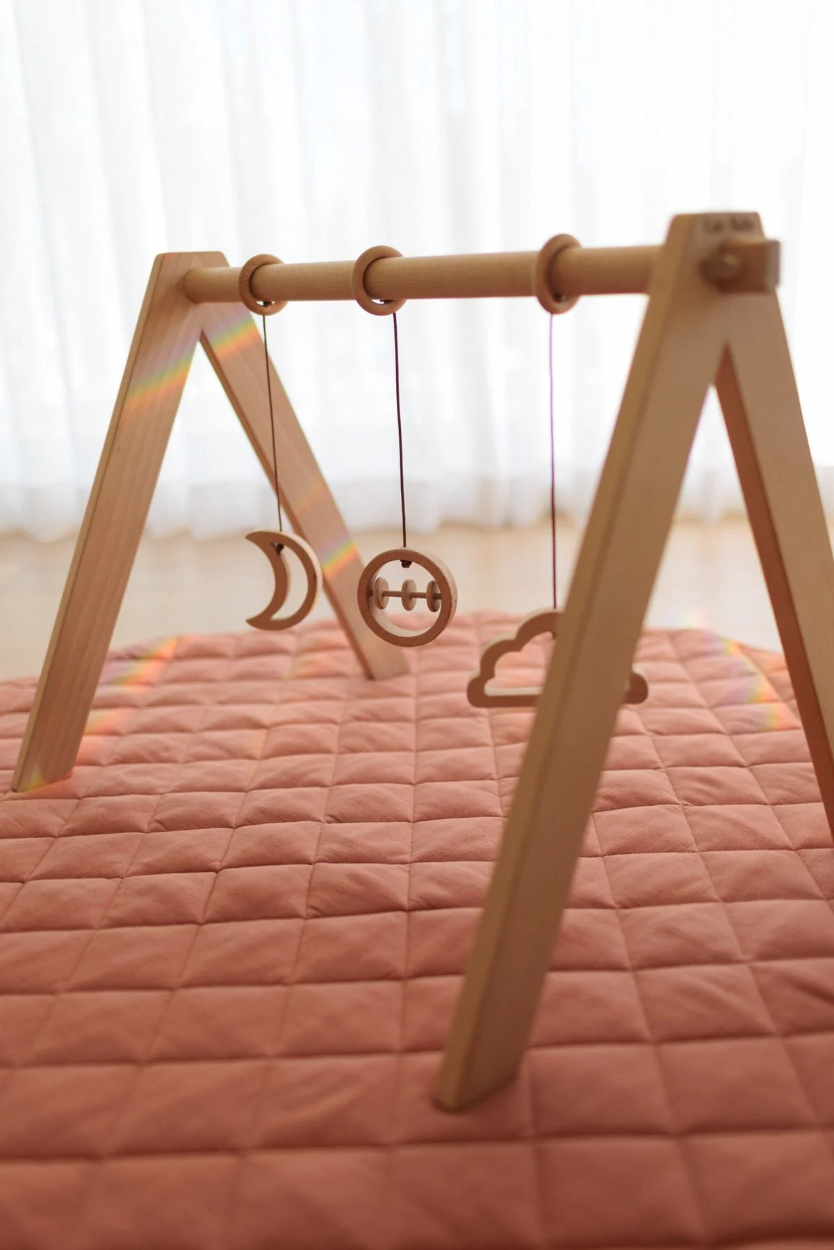 Wooden mobile with moon, cloud, and sunset figures hanging on a pink cushioned surface in front of white curtains.