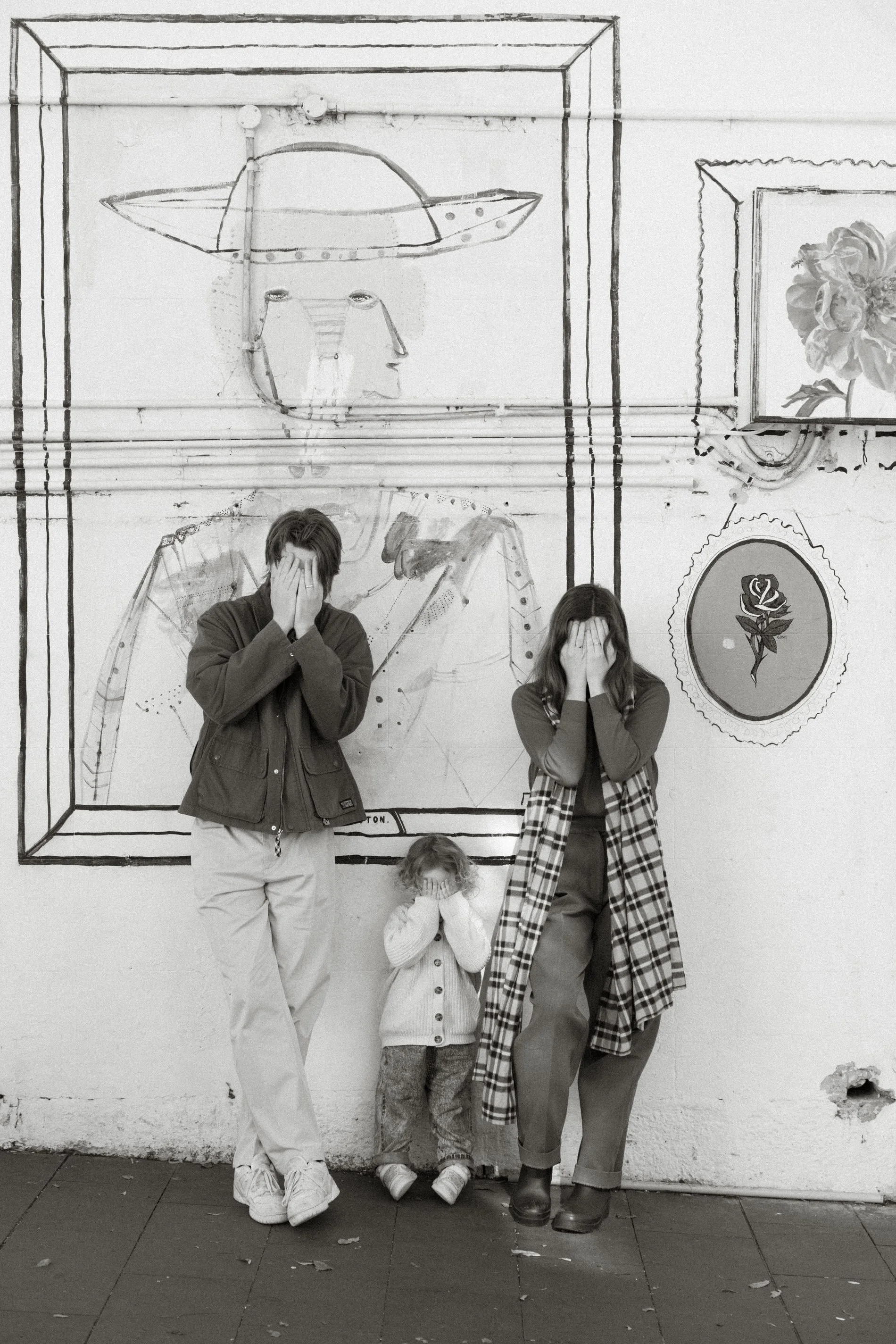 Four individuals, including a young child, standing against a wall with artwork. All four are covering their faces with their hands. The artwork on the wall depicts a stylized face with a hat, a large flower, and other abstract designs.