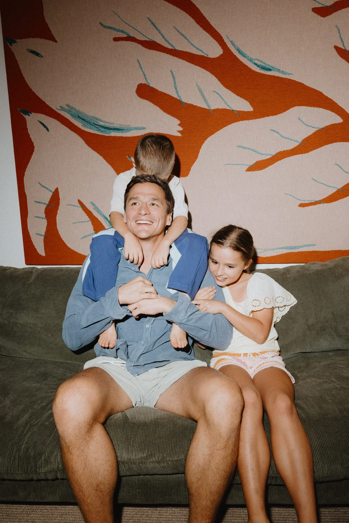 A man sitting on a couch with two children, one girl and one boy. The boy is on the man's shoulders with arms around his neck, and the girl is sitting next to the man, leaning close and smiling. A large abstract painting with orange, light, and teal 