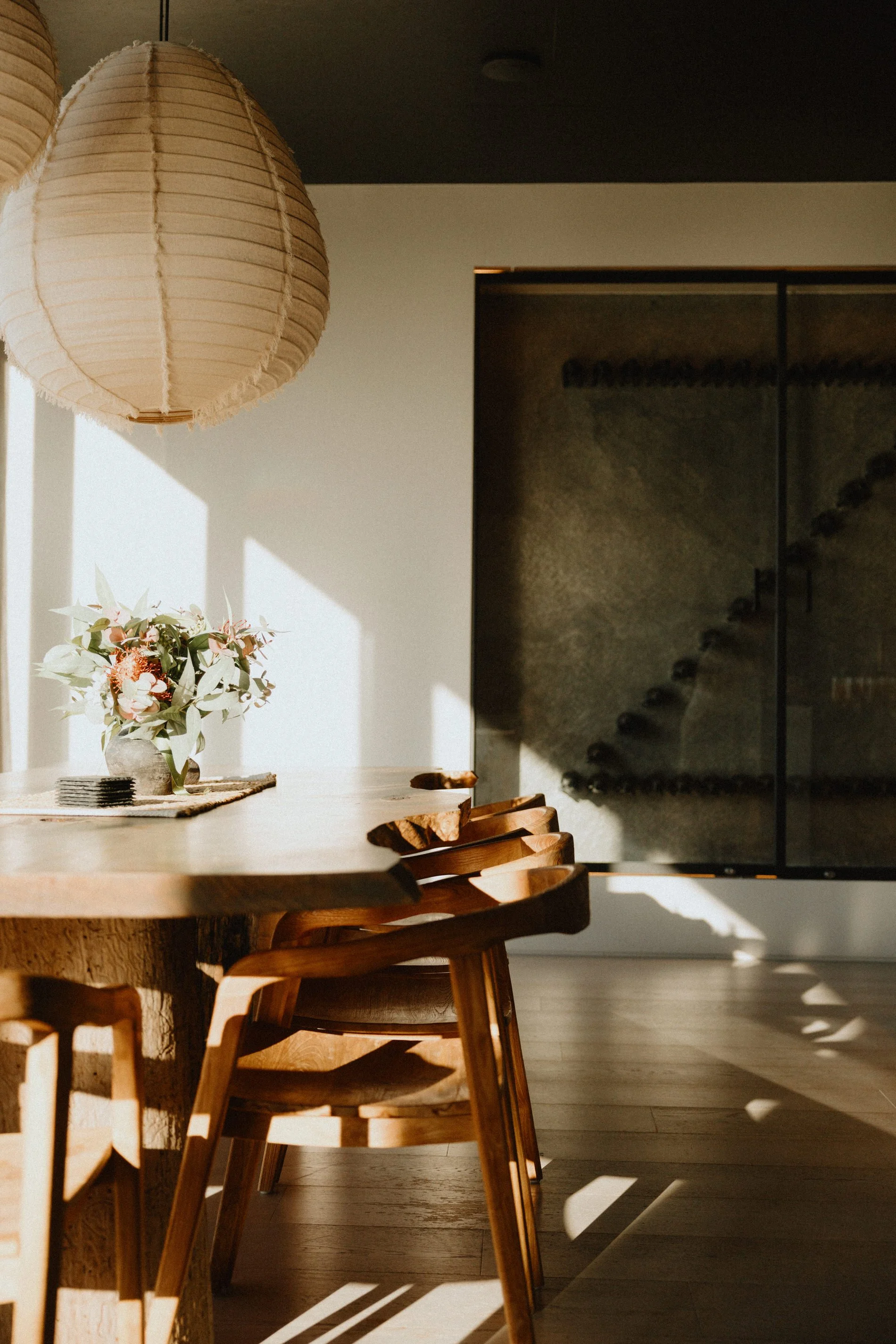 A dining area with a wooden table, surrounded by wooden chairs. A hanging paper lantern provides lighting, and there is a plant with flowers on the table. Shadows cast by sunlight are visible on the floor and walls.