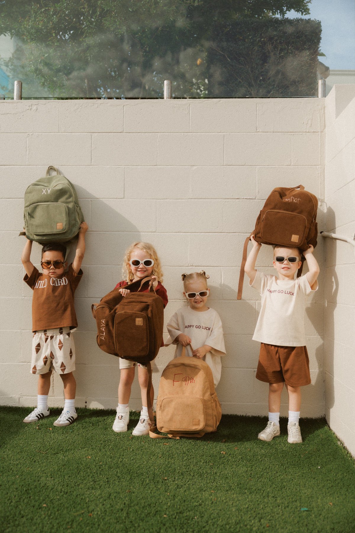 Four children standing against a white wall, wearing sunglasses, with backpacks. The two boys on the ends are holding backpacks on their heads, while the two girls in the middle are holding backpacks. The children are dressed casually, with one boy wearing a brown T-shirt and patterned shorts, and the other boy wearing a white T-shirt and brown shorts. The girls are wearing light-colored dresses. The backpack on the left is green, the girl second from the left is holding a brown one, the girl second from the right has a brown backpack, and the boy on the right has a brown backpack on his head. There is also a light brown backpack with the name “Fawn” written on it on the ground.