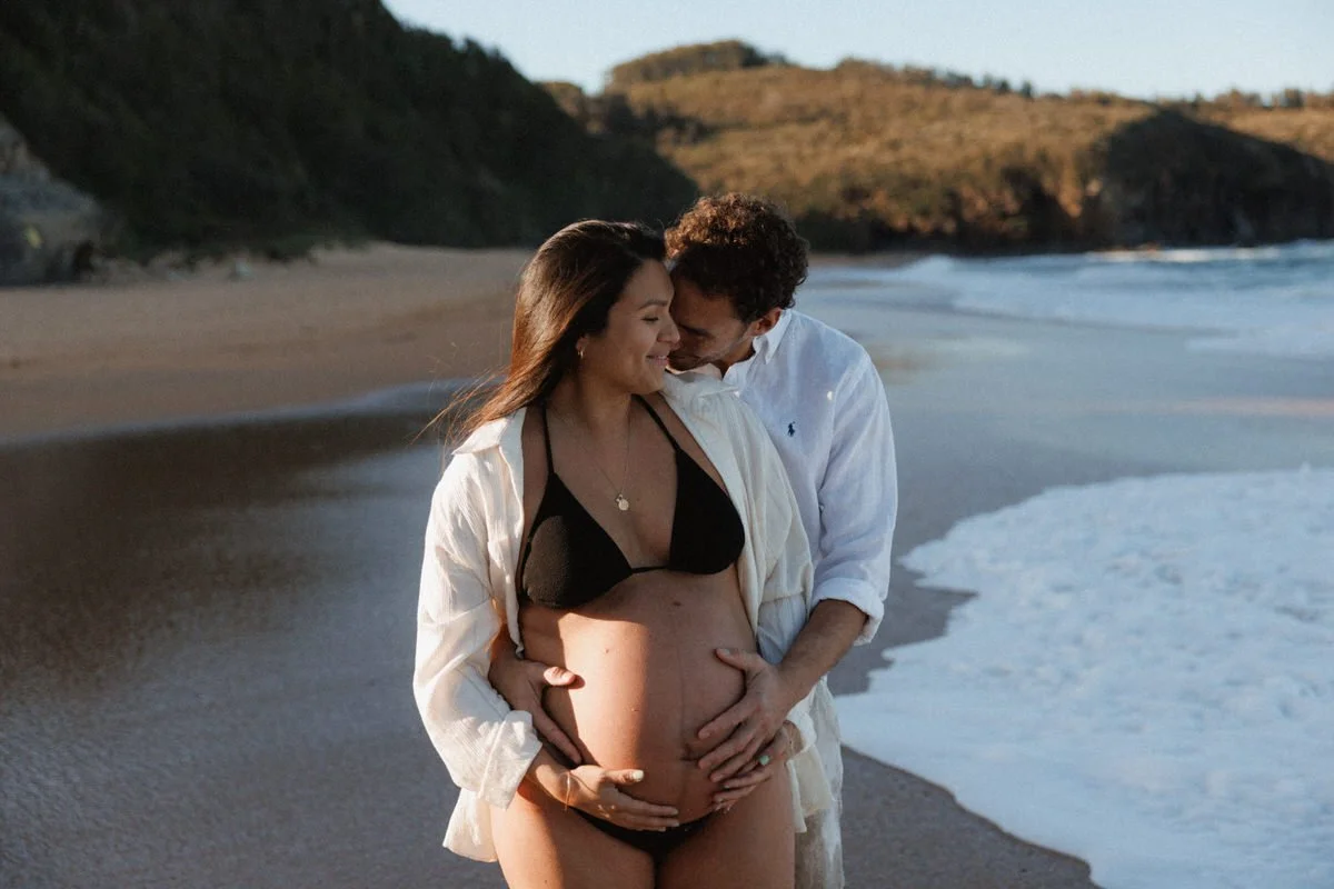 A couple stands on the beach with the woman pregnant, smiling and embracing each other at sunset.