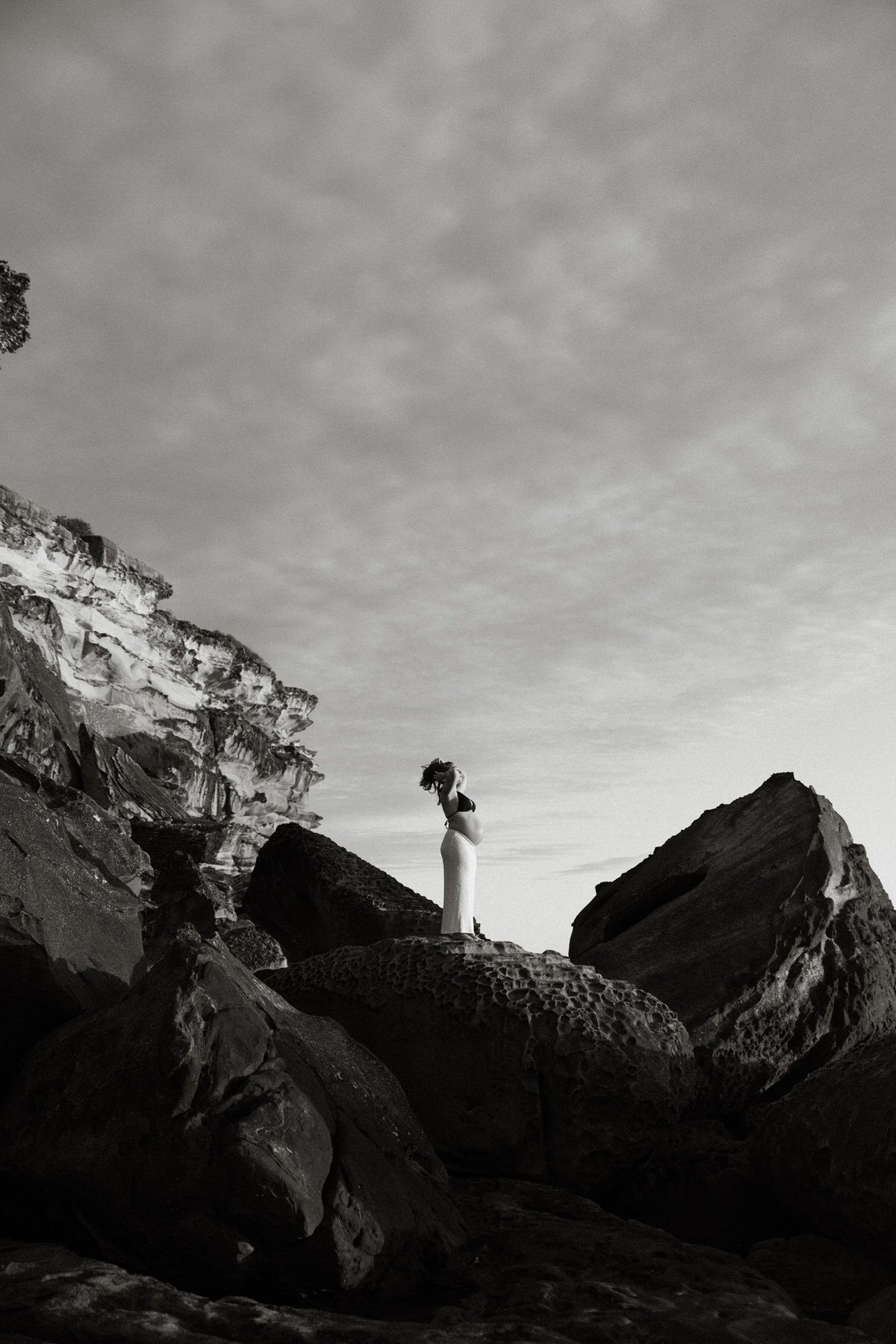 A black and white photograph of a pregnant woman standing on rocks among cliffs, with a cloudy sky above.