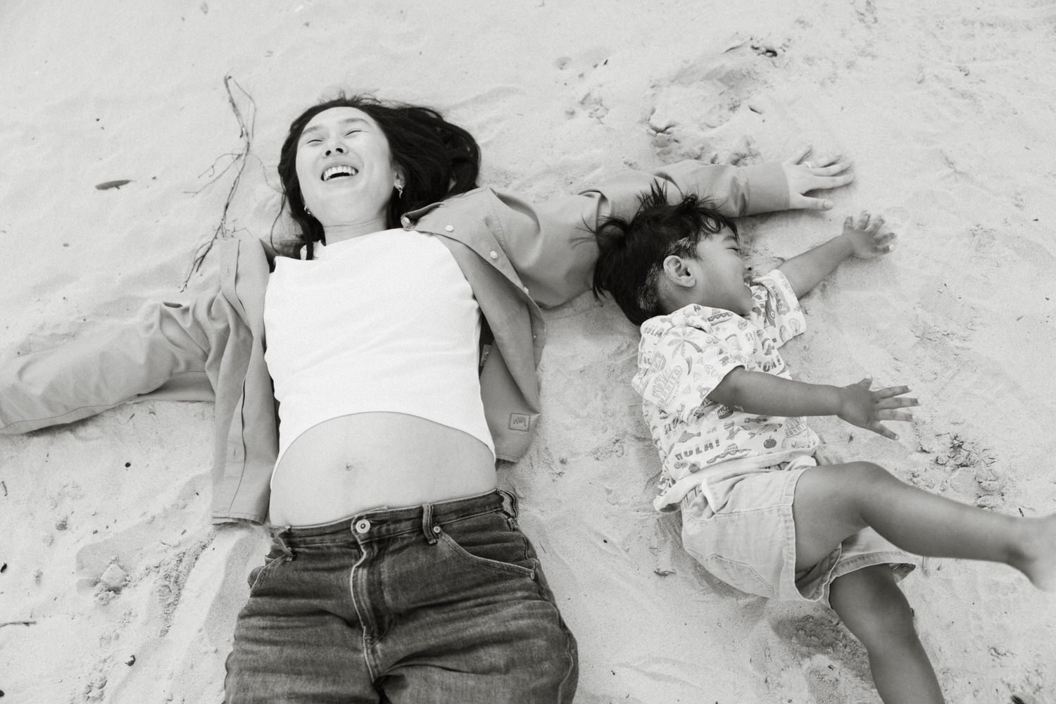 Black and white photograph of a pregnant mother laughing as she lies beside her toddler on the sand during a beach maternity session.
