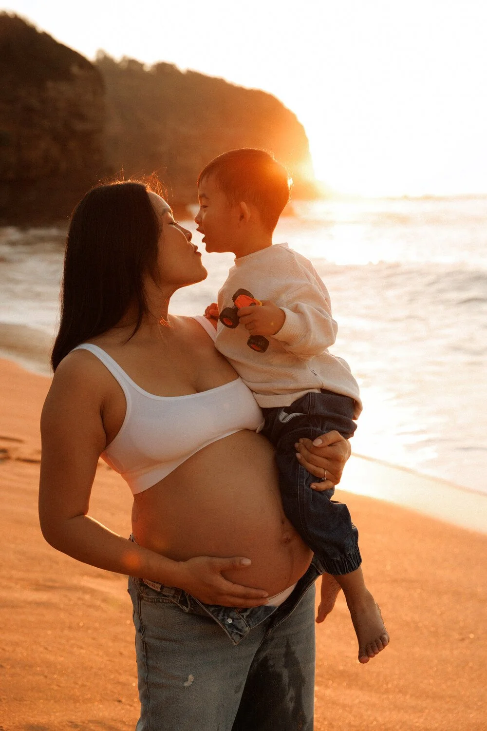 Pregnant mother holding her toddler as they lean in for a kiss beside the sunlit ocean.
