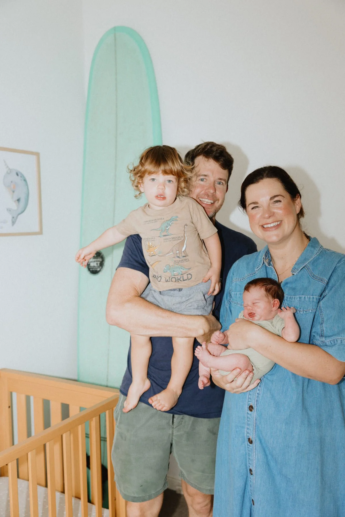 A family of four, including a man, a woman, a young boy, and a newborn baby, standing together in a nursery, with a pastel green surfboard painting on the wall behind them.