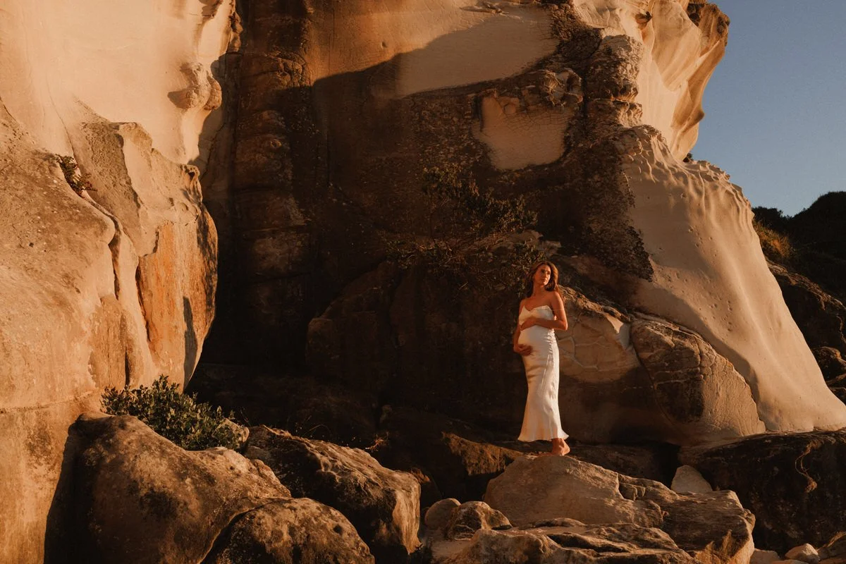 A woman in a white dress standing on rocks in a desert landscape with large sandstone formations, during golden hour.