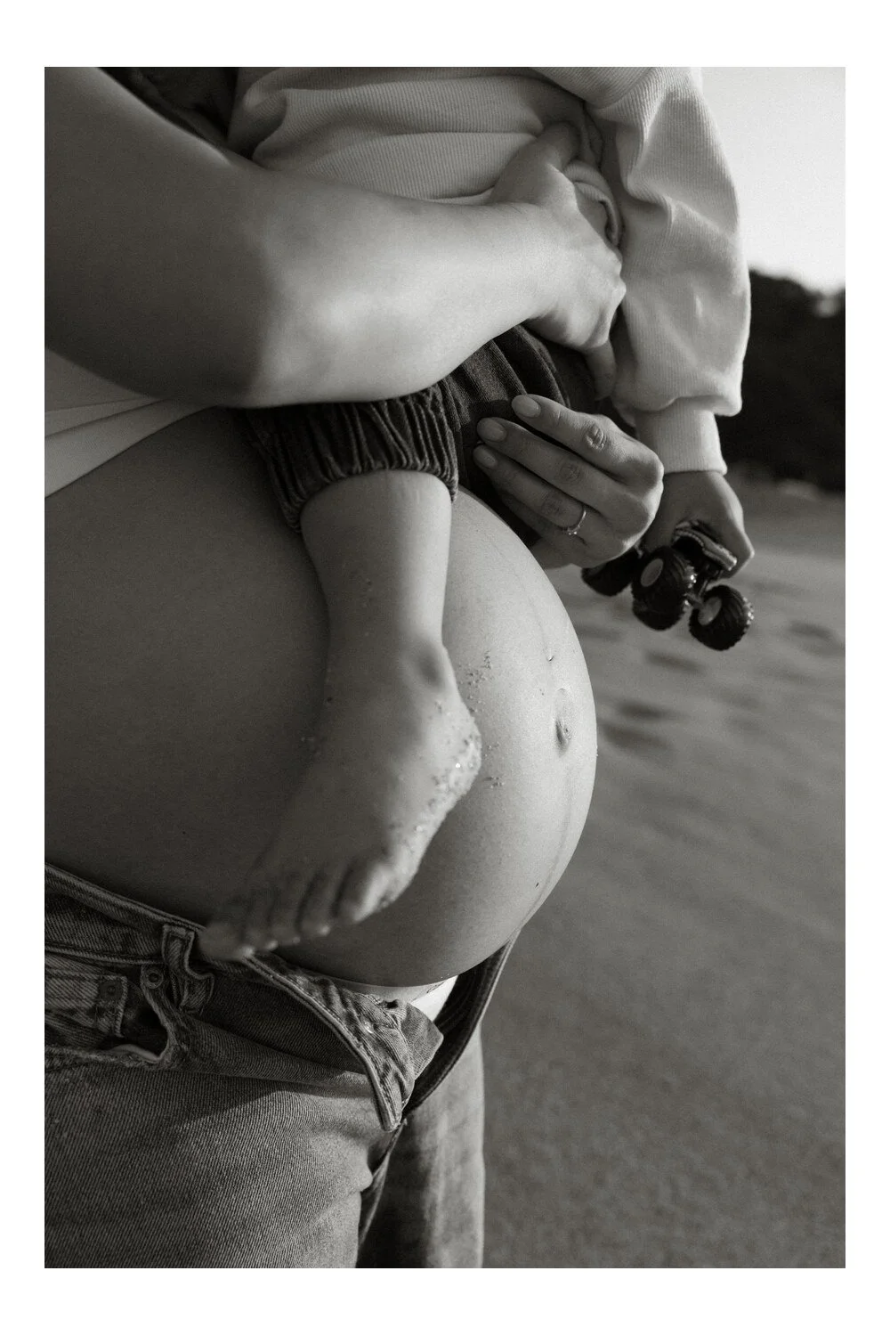 Black-and-white close-up of a toddler’s sandy foot resting against their mother’s baby bump.
