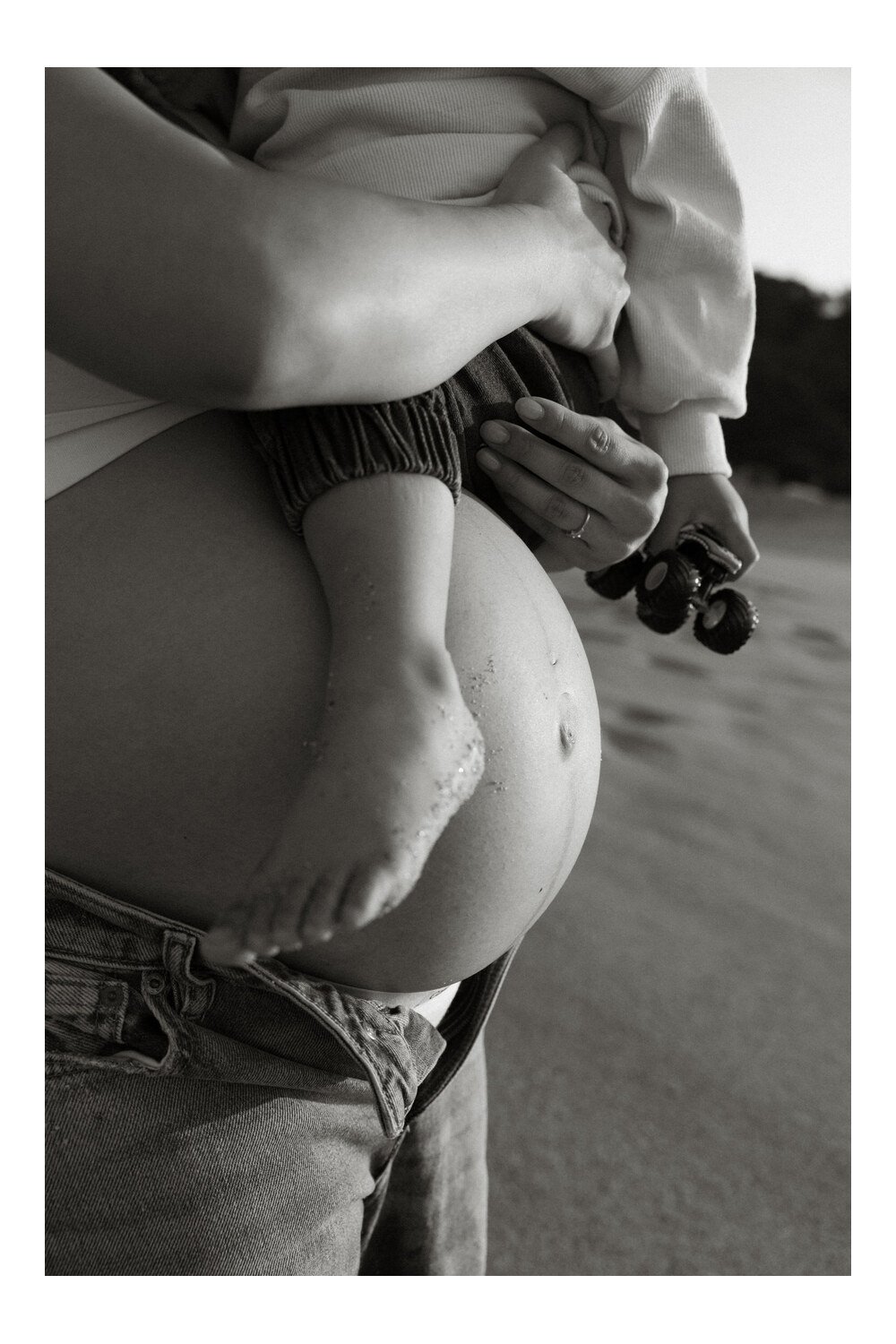 Black-and-white close-up of a toddler’s sandy foot resting against their mother’s baby bump.