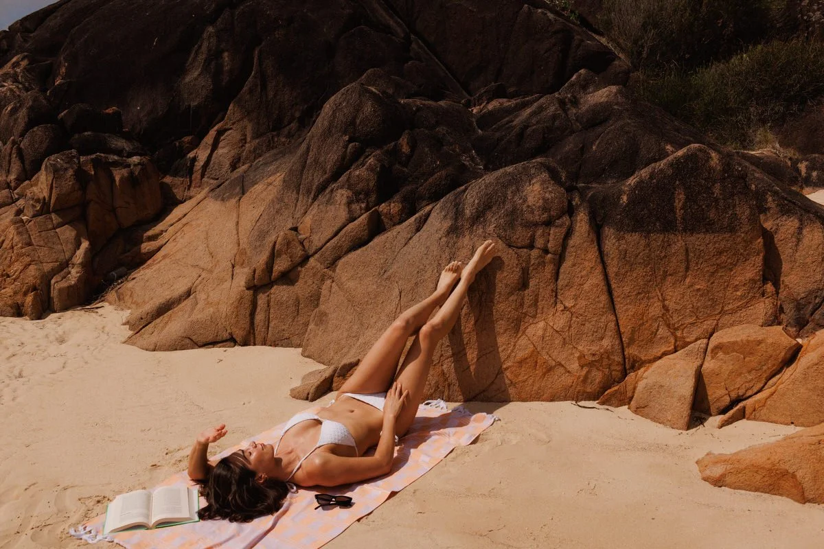 Woman lounging on a beach towel by rocks, wearing a white bikini, with an open book and sunglasses nearby.