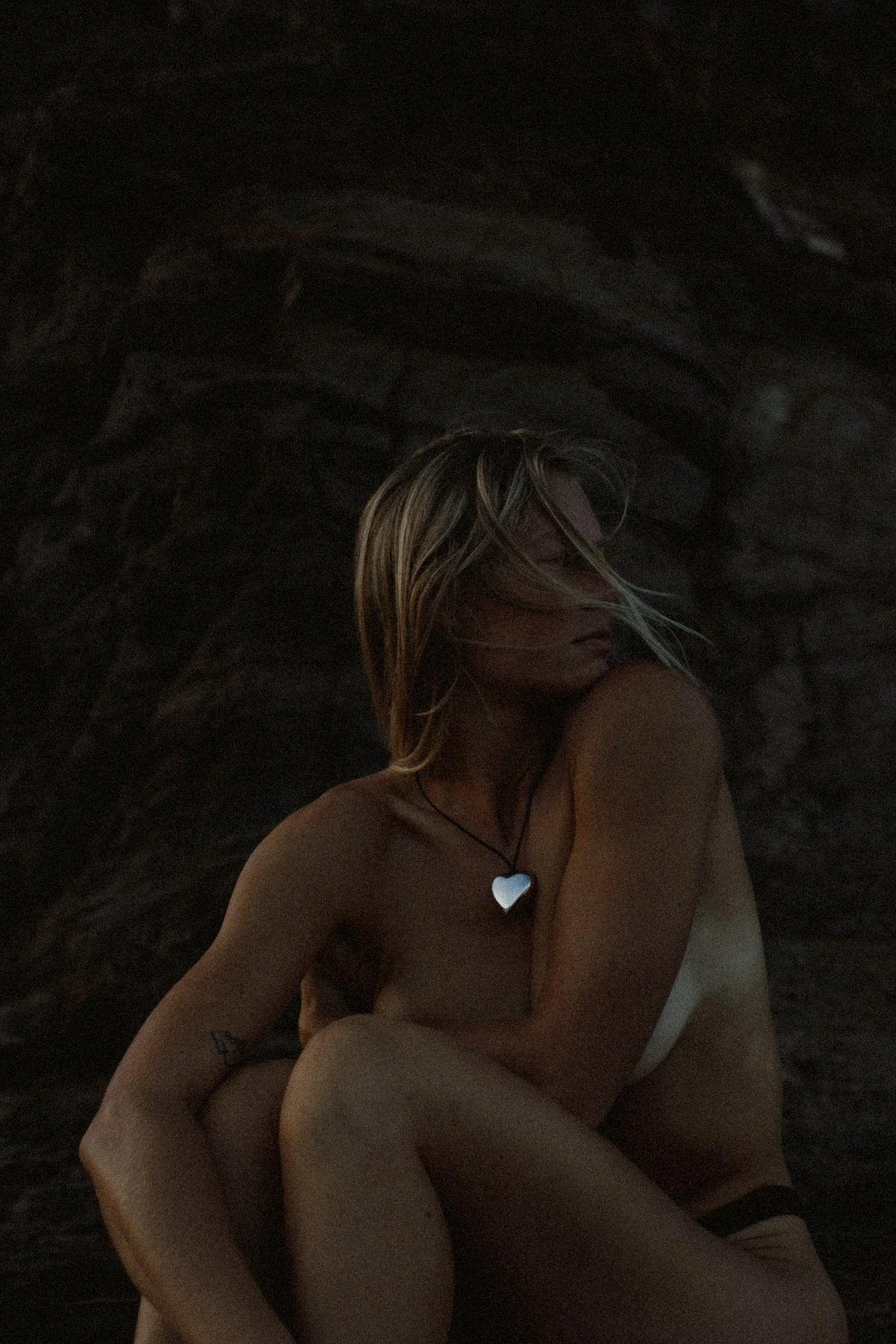 A woman with blonde hair and a heart-shaped necklace sitting against a dark, rocky background, with her arms wrapped around her knees.