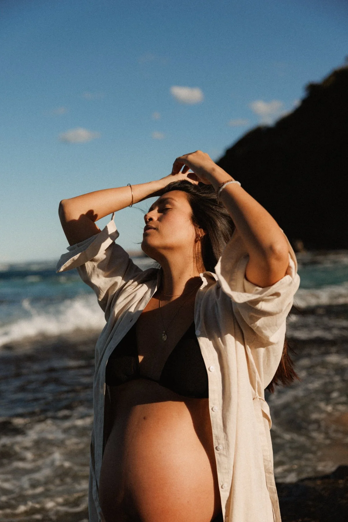 Young woman standing on the beach with her eyes closed and hands on her head during sunset, wearing a black bra and light-colored shirt, with ocean waves and cliffs in the background.