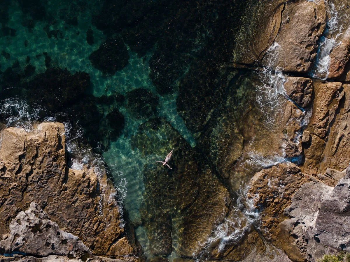 A person swimming in a clear, rocky coastal waters with large rocks surrounding the area.