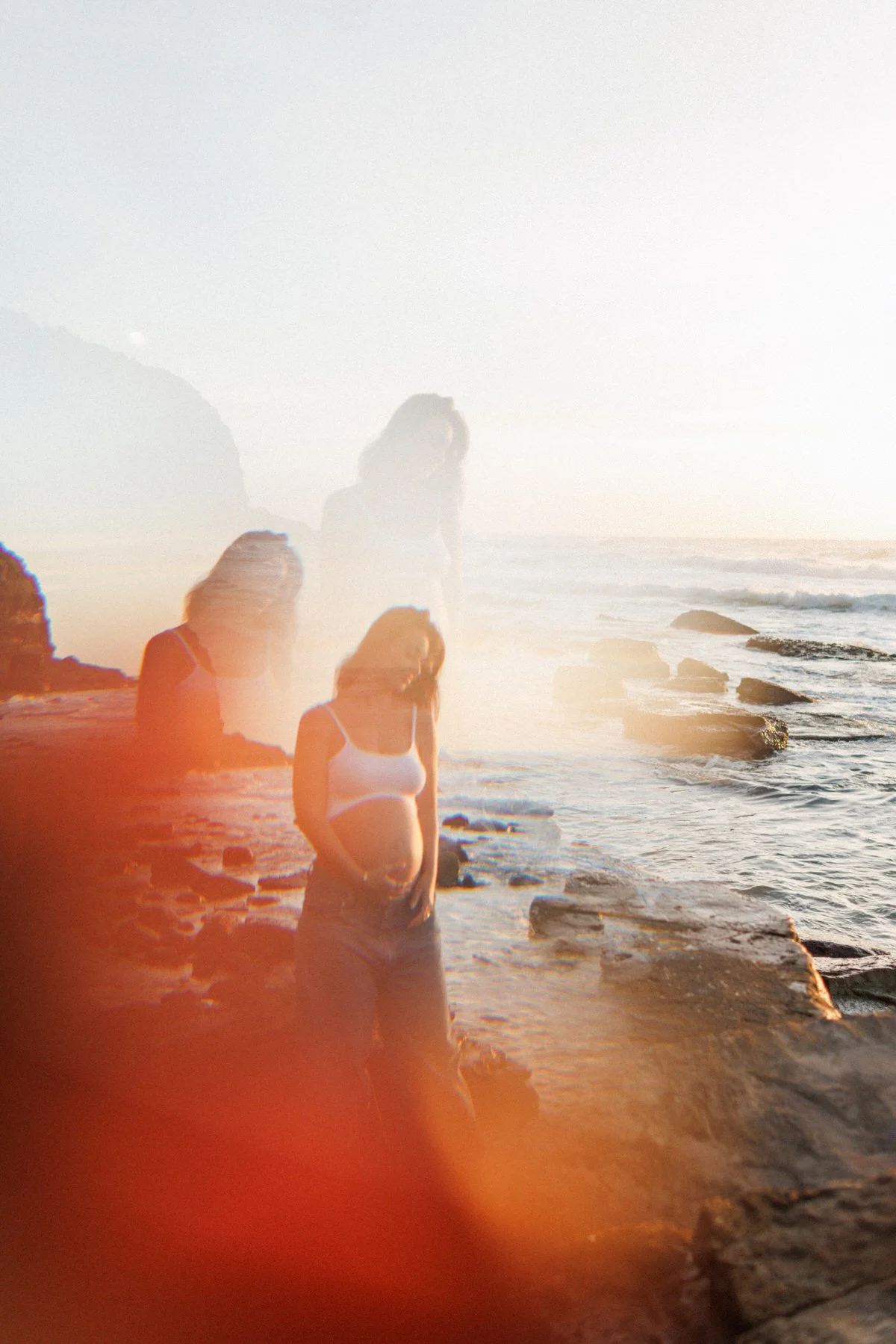 A woman in a white tank top and shorts standing on a rocky beach during sunset with ocean waves in the background.