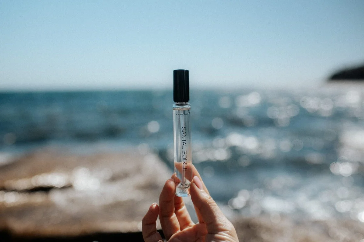 Person holding a small bottle of perfume labeled 'Santal Sourire' with a beach and ocean in the background.