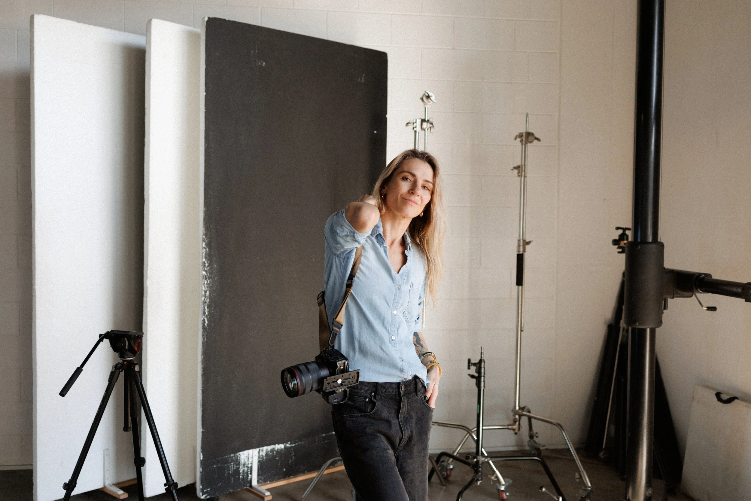 A woman with shoulder-length blonde hair wearing a blue shirt and black pants, standing in a photography studio surrounded by photography equipment including tripods, softboxes, and a camera. She is smiling at the camera with a black backdrop behind 