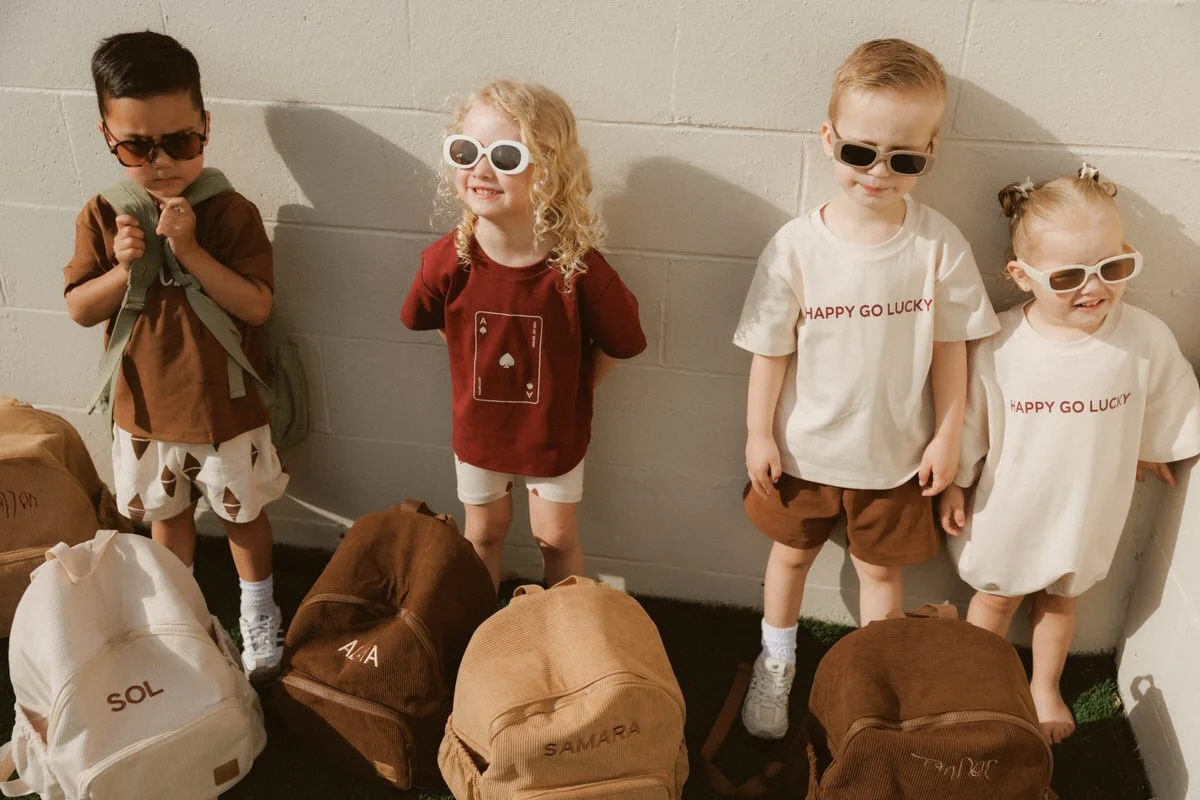 Four young children standing against a white brick wall, wearing sunglasses, with backpacks on the ground in front of them.