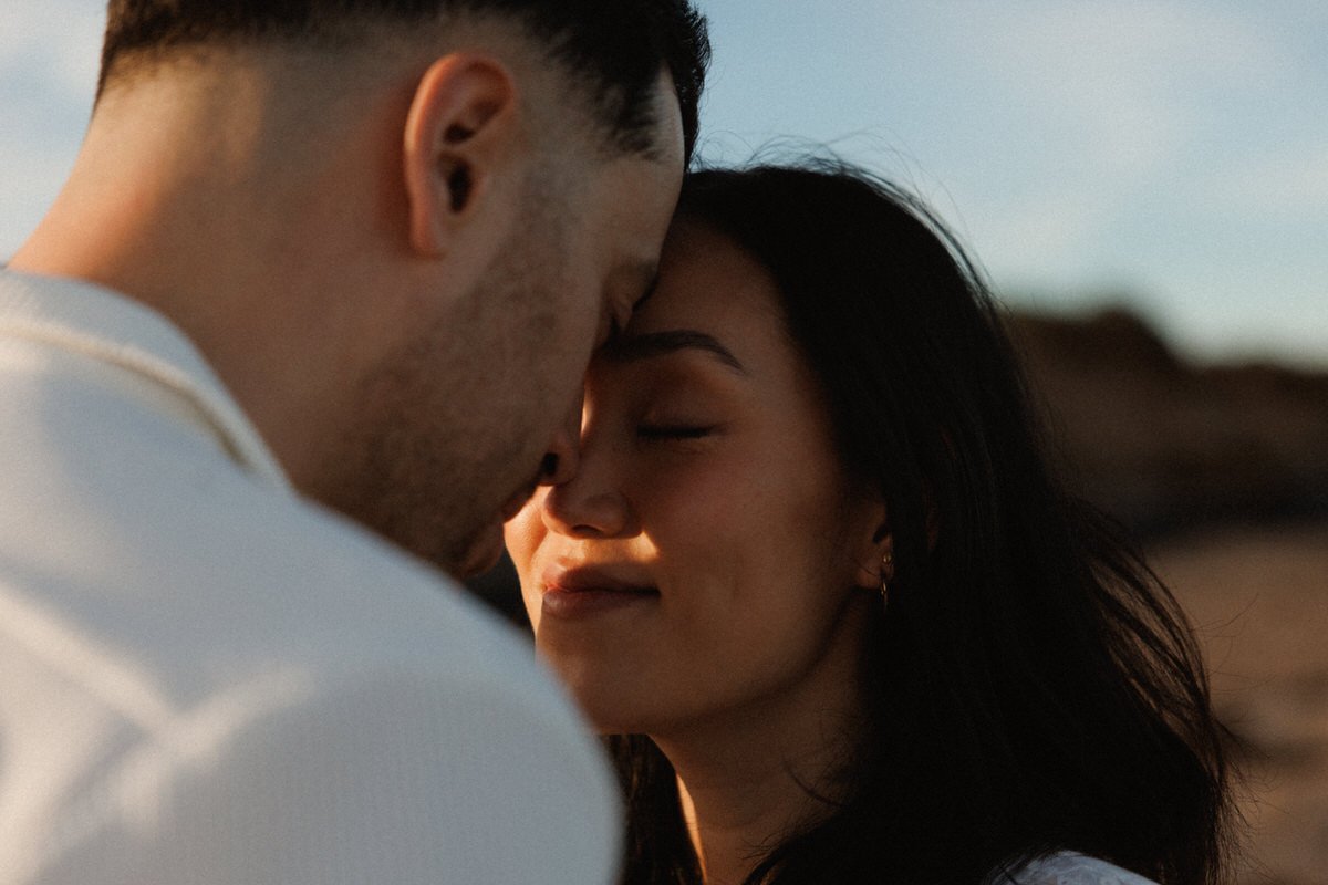 A close-up of a couple with their foreheads touching, eyes closed, sharing an intimate moment outdoors during sunset.