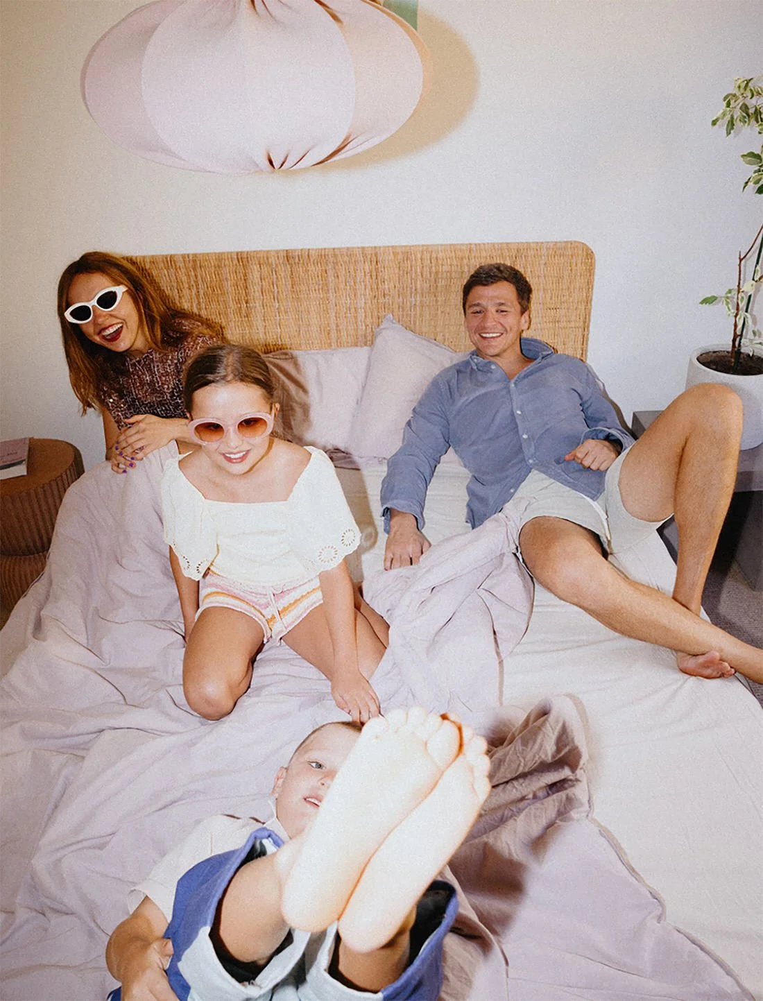 A family of five on a bed, smiling and having fun, with some members wearing sunglasses; a woman with red hair and sunglasses, a man in a blue shirt, two girls in white tops and sunglasses, and a boy in shorts with his feet close to the camera.