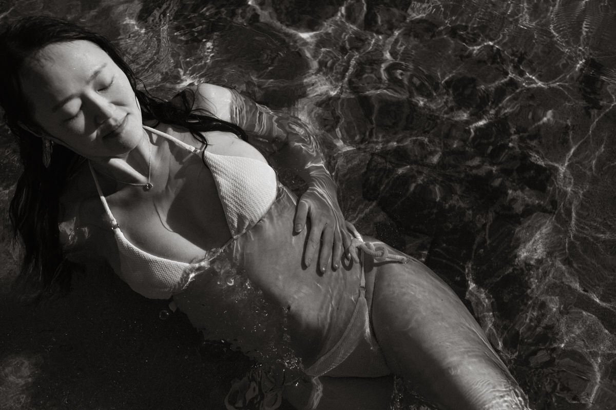A woman with long dark hair floating in water, wearing a bikini, with her eyes closed and hand resting on her stomach.