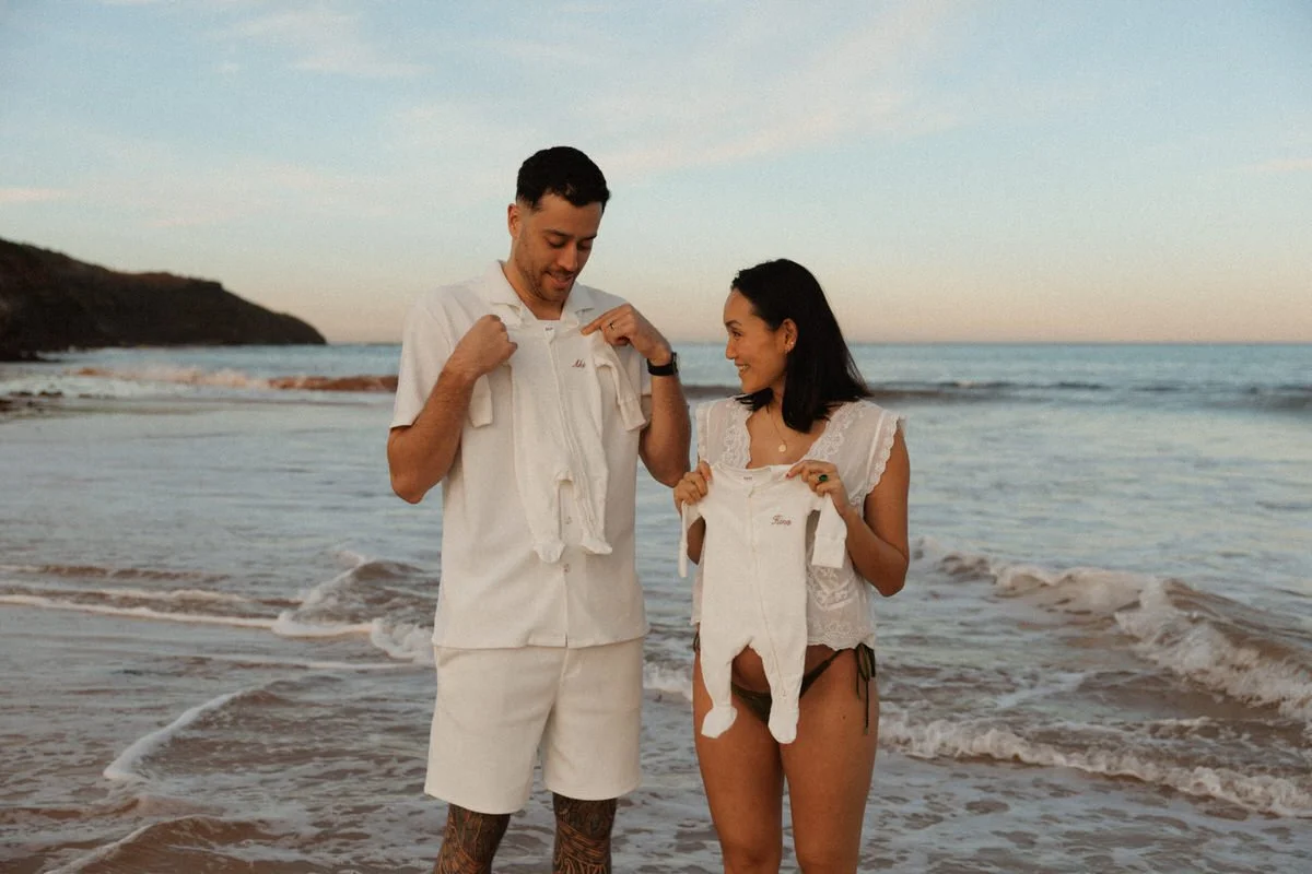 A man and woman on the beach holding matching white outfits, smiling at each other during sunset.