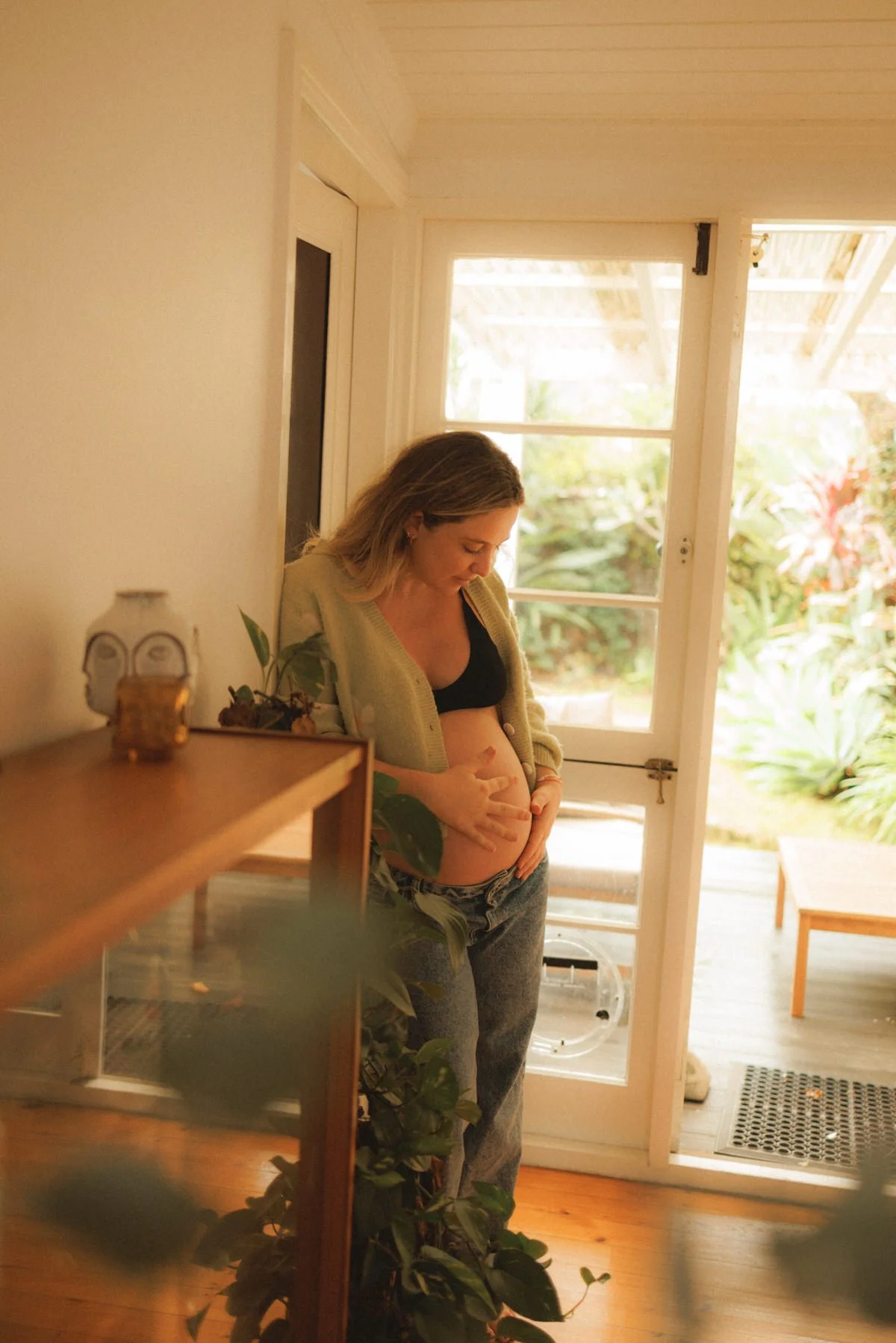 Pregnant woman standing near a door with large windows, gently touching her belly and looking down, in a cozy home with plants and wooden furniture.