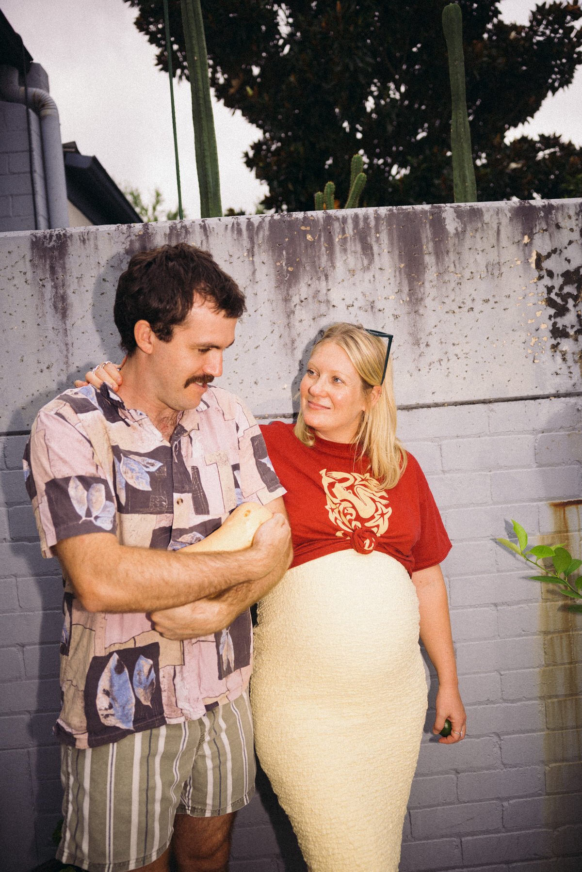A man with dark hair and a mustache wearing a patterned short-sleeve shirt and striped shorts is holding a ball of bread, standing next to a pregnant woman with blonde hair wearing a red T-shirt with a dragon print and a cream-colored knitted dress, 