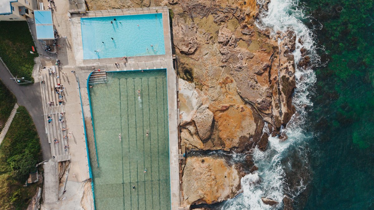 Bird's eye view of a swimming pool and smaller pool next to rocky shoreline with waves crashing against rocks.