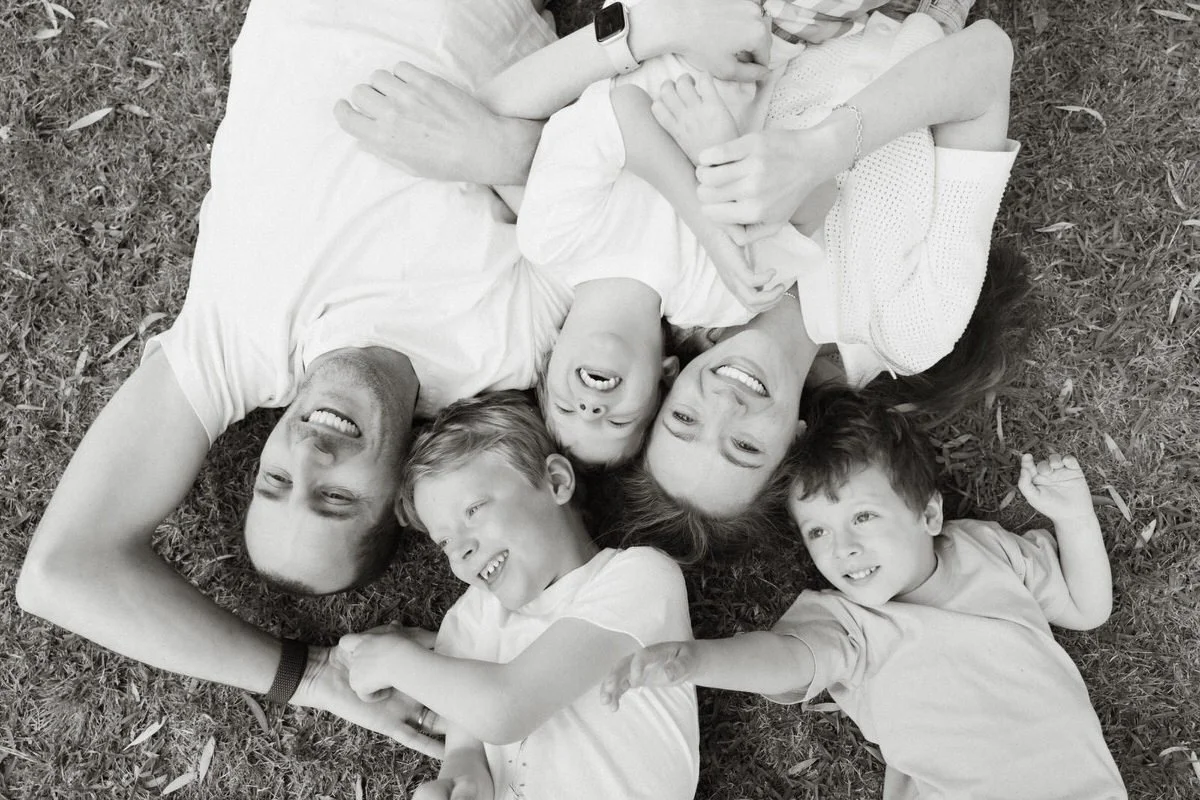 A family of six lying on grass, smiling, with the parents and four children all looking up and laughing.
