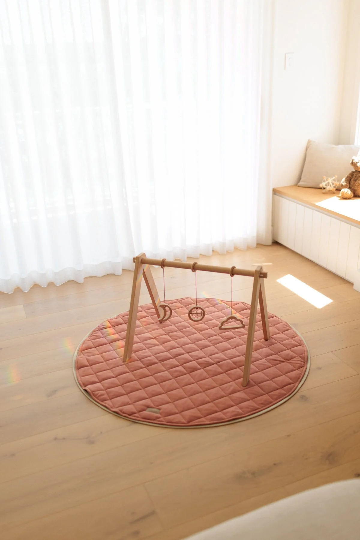 A children's wooden play gym with hanging toys, placed on a pink circular rug in a bright room with white curtains and a bench with pillows and stuffed animals.