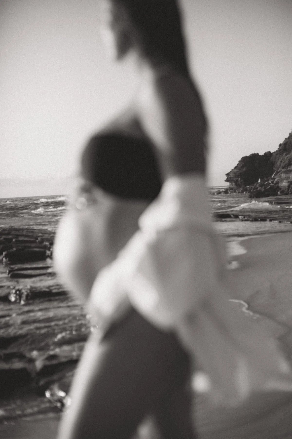 Blurred black-and-white photo of a woman and child at the beach, with ocean waves and a rocky cliff in the background.